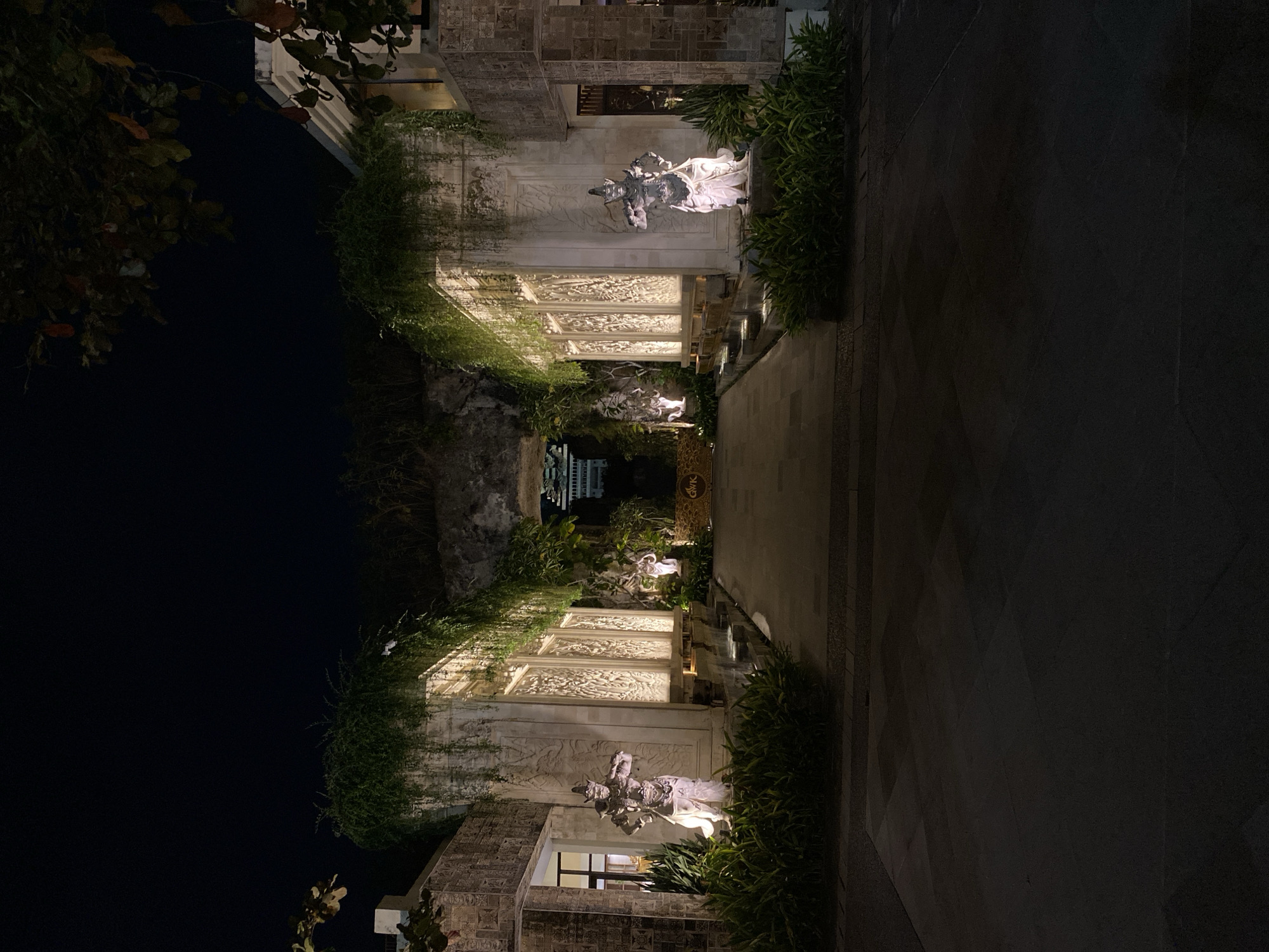 This media file depicts an outdoor scene at night, possibly in Kuta Selatan, Indonesia, given the architectural and decorative style. The foreground features a pathway made of stone tiles, bordered by lush green foliage. Beyond the pathway, there's an illuminated walkway leading to what appears to be a building with intricate stone carvings on its facade.
Dominating the scene are two statues, one larger and closer to the viewer, and another smaller one further in the distance. Both statues appear to depict figures in a dynamic pose, possibly mythological or historical characters, rendered in a light-colored material, likely stone. The statues and the carved walls are artfully lit, creating dramatic shadows and highlighting the detailed craftsmanship.
The overall atmosphere is serene and somewhat mysterious due to the nighttime setting and the dramatic lighting. The presence of dense greenery suggests a tropical or subtropical environment, consistent with the Indonesia location context. The architectural style, with its ornate carvings and stone elements, hints at a culturally rich location, perhaps a temple, a resort, or a high-end establishment.
The image is oriented vertically, with the top of the frame showing a dark sky and some foliage from a tree. The lighting suggests artificial illumination, implying that the scene is accessible and perhaps a destination for visitors to experience at night. There are no discernible activities or events taking place; the focus is on the static beauty of the architecture, the statues, and the interplay of light and shadow. A golden sign with text is partially visible on the right side of the image, but the text itself is too blurry to be read.