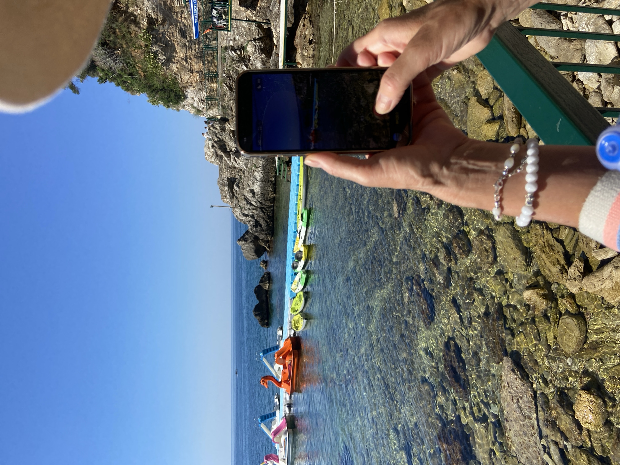 The image depicts a sunlit scene at a coastal location, likely in Budva, Montenegro, as suggested by its characteristic rocky coastline and clear waters. In the foreground, a person's hands hold a smartphone, capturing a view of the water. The phone screen displays a photograph of several kayaks and pedal boats floating on the sea.
The water is exceptionally clear, revealing a rocky seabed with various stones and pebbles. A series of colorful kayaks, in shades of yellow, green, and white, are lined up on a blue floating platform. Adjacent to the kayaks, there are two pedal boats: one shaped like a pink unicorn and another orange swan-shaped boat with a slide.
In the background, a rocky cliff face rises steeply, covered with vegetation. Several people are visible on a path winding along the cliff. A small sign on a building at the top of the cliff reads "WATER SPORT CLUB". The sky is a bright, cloudless blue, and the sea stretches out to the horizon, with a small boat visible in the distance. The overall impression is one of a recreational beach area on a clear, warm day.