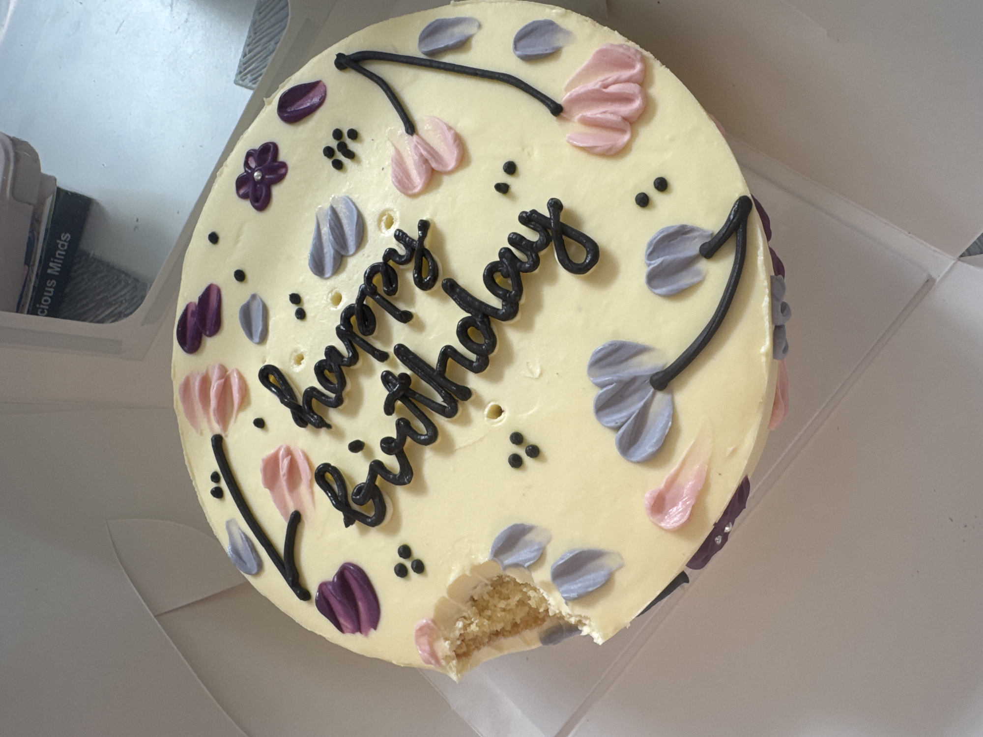 A round birthday cake with light yellow frosting is prominently featured, slightly angled from an overhead perspective, resting inside a white cardboard box. The cake is intricately decorated with various frosting elements: "happy birthday" is written in elegant black script across the center. Around the text, there are stylized floral or leaf designs made from pastel-colored frosting in shades of pink, light grey-purple, and a darker, richer purple. Small clusters of black frosting dots are scattered across the cake's surface, adding to the decorative pattern.
A noticeable bite has been taken from the bottom right side of the cake, revealing the light, crumbly texture of the cake layers beneath the frosting. The edges of the cake are neatly frosted, with some of the purple and pink decorations extending around the sides.
The white cake box is partially open, with its clean, sharp edges visible around the cake. In the upper left corner of the image, a hint of another object, possibly a book or packaging, can be seen, with the text "cious Minds" partially legible on its side. The overall lighting is bright and even, suggesting an indoor setting. The scene captures the immediate aftermath of someone enjoying a slice of a beautifully decorated birthday cake.