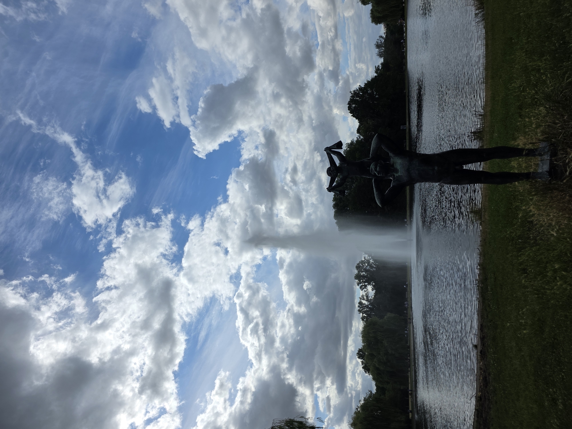 The image depicts a view of the Fontänendenkmal in Halle (Saale), Germany. In the foreground, a bronze sculpture of two figures, a larger one supporting a smaller one on its shoulders, stands silhouetted against a tranquil lake. The larger figure is nude and stands with its legs slightly apart, while the smaller figure, also nude, is perched on its shoulders, holding its arms raised. Behind the sculpture, the lake stretches out, its surface rippled by a gentle breeze. A tall, powerful fountain erupts in the center of the lake, sending a plume of water high into the sky. The background is filled with lush green trees, creating a natural barrier between the lake and the sky. The sky above is a dramatic canvas of blue, punctuated by a mixture of dark, billowing clouds and wispy cirrus formations, suggesting dynamic weather conditions. The overall scene conveys a sense of serene beauty and dramatic natural elements.