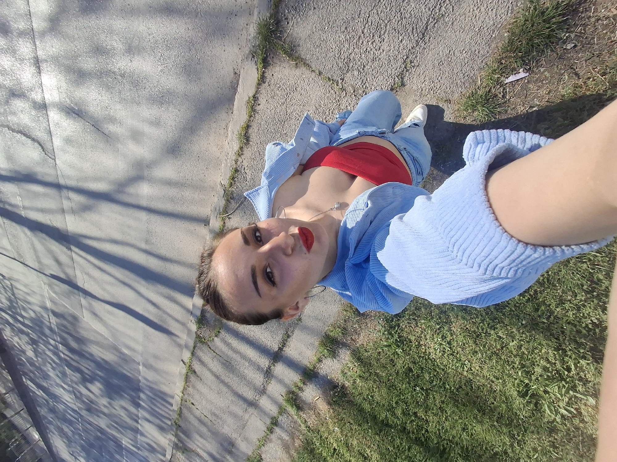 A young woman is taking a selfie outdoors. She is wearing jeans, a red top, and a light blue corduroy jacket. She is wearing red lipstick. The background is a sidewalk and grass. The location is not identifiable as Sofia, Bulgaria from visual elements alone.