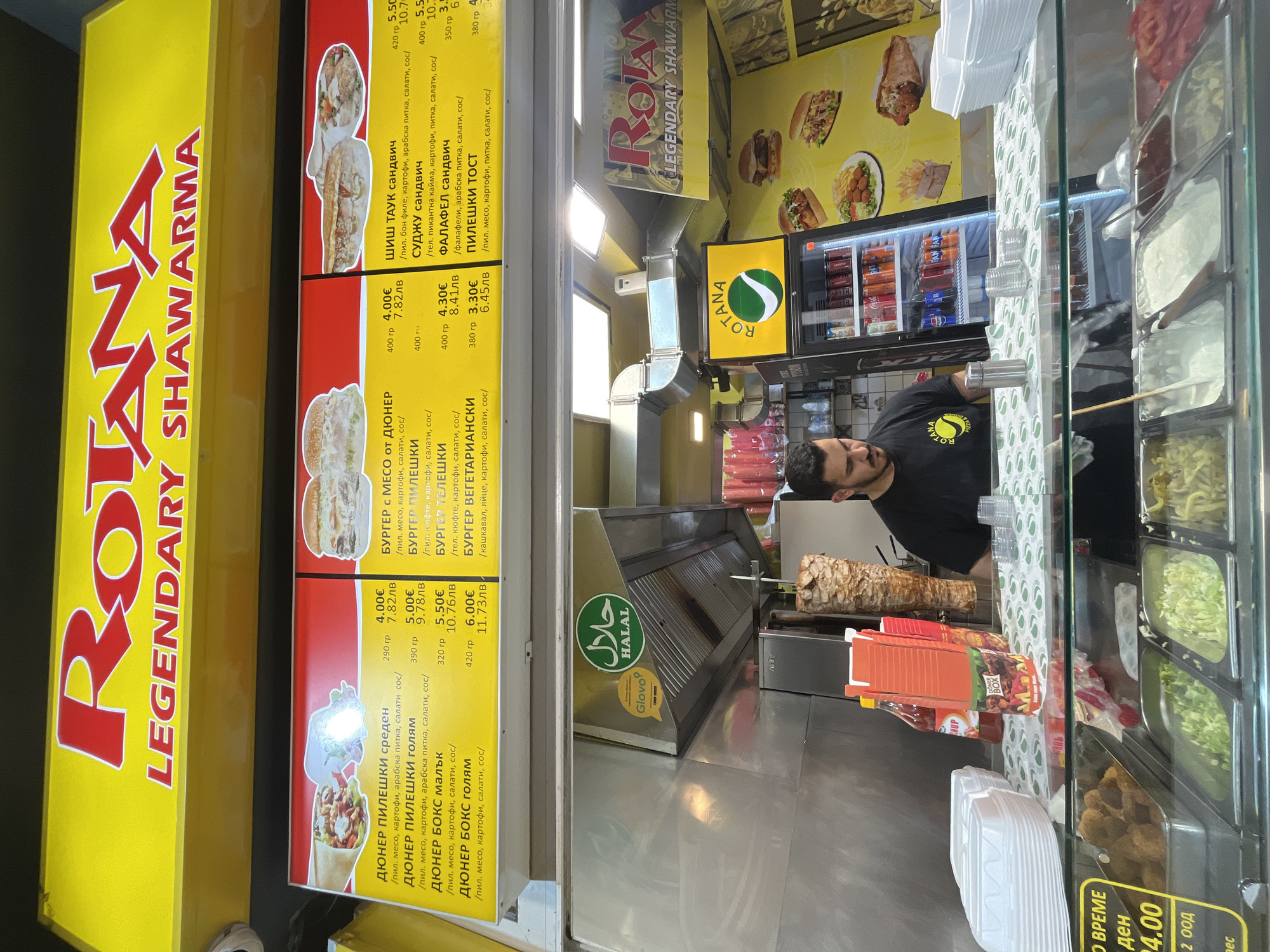 Here's a detailed description of the image:
**Scene:** The image captures the interior of a small fast-food restaurant in Sofia, Bulgaria, specializing in shawarma and related items. The focus is on the service counter and the food preparation area behind it.
**People:** A man with dark hair and a beard is visible behind the counter. He is wearing a black t-shirt with the brand logo "ROTANA" and appears to be attending to the food preparation.
**Objects:**
* Food preparation equipment: Shawarma meat is spitted and ready to be sliced, a grill, and serving containers with fresh ingredients for salads and sandwiches.
* Menu boards: These are placed prominently above the counter and display the restaurant's offerings in Bulgarian, including various shawarma types ("дюнер"), burgers, and sandwiches, along with their prices in both Euro and Bulgarian Lev currency.
* Drinks refrigerator: A refrigerator is stocked with various beverages, including sodas.
* Restaurant branding: The "ROTANA" logo is present on the menu boards, the man's t-shirt, and other signage.
* Delivery Service logo: The "Glovo" logo on the grill equipment.
* Halal symbol: The Halal symbol indicates that the establishment adheres to Halal dietary standards.
**Actions and Interactions:** The man is working in the food preparation area, likely preparing or serving a customer's order.
**Environment:** The environment is clean and functional. The stainless steel surfaces of the food preparation area are well maintained. The overhead lighting is bright. The background shows other menu displays and the drinks refrigerator.
**Overall Impression:** This is a shot of a busy shawarma and fast food restaurant in Sofia, captured during what seems to be an active service period. The signage, branding, and menu are designed to be visually appealing and informative to customers.