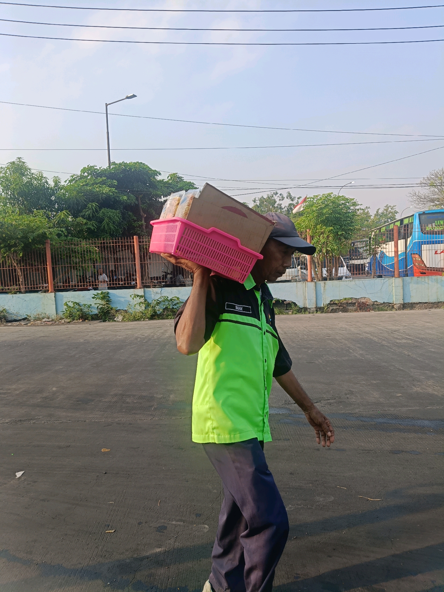A man wearing a bright green and black collared shirt with the name "IMAM" visible on the left chest, dark pants, and a dark baseball cap walks from right to left across a paved surface. He is carrying a pink plastic basket filled with goods, balanced on his left shoulder, with a cardboard box resting on top of the basket. The man's right arm is bent at the elbow, and his hand is held slightly away from his body. In the background, there is a metal fence, green foliage, a light pole, and a blue and white bus. The environment appears to be an outdoor urban setting, possibly near a road or parking area. The sky is visible, with a few thin clouds.