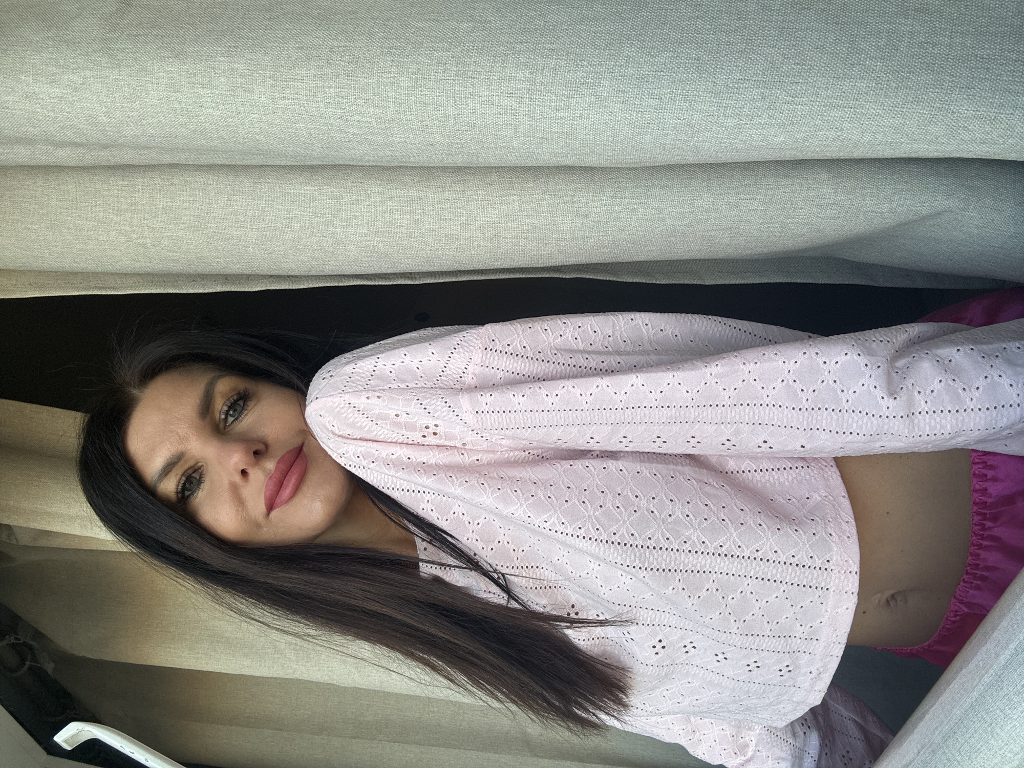 A young woman with long, dark hair is captured in a close-up, rotated portrait-style image. She is looking directly at the viewer with a calm expression, her lips painted with a muted pink lipstick. Her eyes are a light color, possibly green or hazel, accentuated with dark eyeliner and mascara. She wears a light pink, long-sleeved crop top made of eyelet lace fabric, revealing a sliver of her midriff and navel. Bright pink, possibly satin, shorts or pants are visible below the crop top.
The woman appears to be positioned within an interior space, likely leaning or sitting against light-colored, possibly beige or light grey, fabric panels that resemble curtains, blinds, or upholstery. These panels form a backdrop and partially enclose her. A darker area is visible behind her head and shoulders, creating a contrast. In the bottom left corner, a white, curved handle or fixture is partially visible, further suggesting an indoor setting, possibly within a vehicle or room with window coverings. The overall lighting is soft and even, indicating daytime. No visible text is present. The location context is Chaikovsky, Russia.