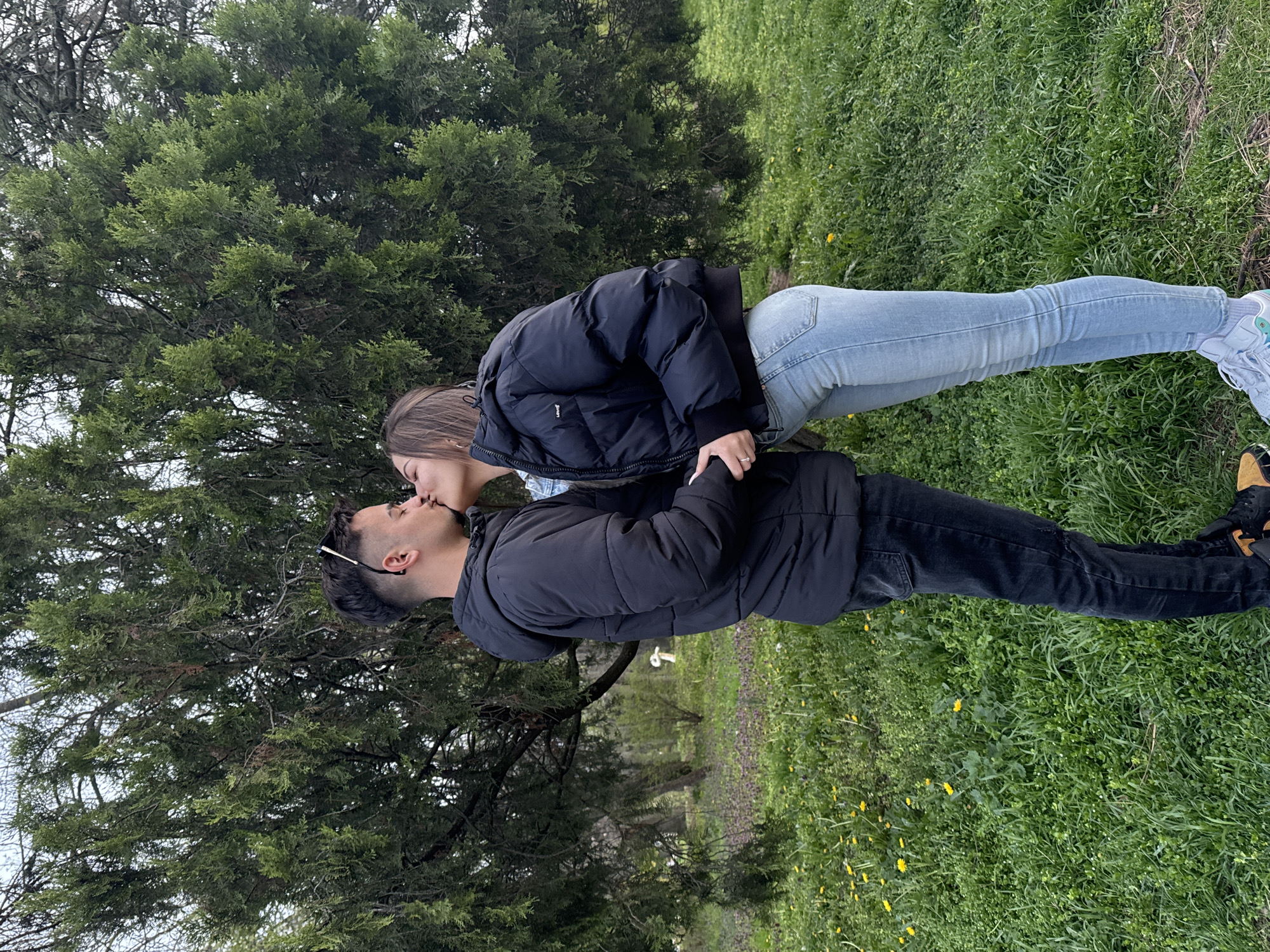A young couple is kissing in a park setting. The woman, wearing a dark puffer jacket and light blue jeans, is standing to the right of the man. The man, wearing a black puffer jacket and dark jeans, is standing to the left. They are holding hands. A large tree is directly behind them. The ground is covered in green grass and small yellow flowers. The location is likely a park in Sofia, Bulgaria.