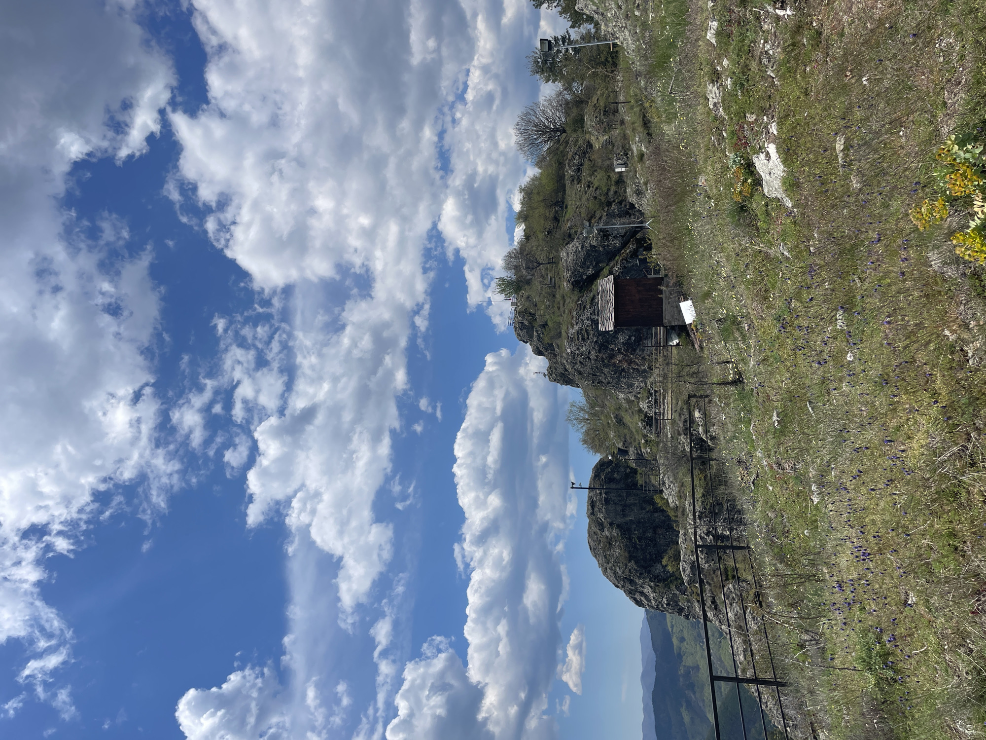 The image depicts a view from a hillside in Gradat, Bulgaria. The right side of the frame shows a grassy, rocky slope dotted with purple flowers. A black metal fence runs along the edge of the cliff. Further up the slope, there's a small wooden structure or shack attached to the rock face, and some supporting framework is visible nearby. The left side of the frame is dominated by a bright blue sky with fluffy white clouds. A distant mountain range is faintly visible at the bottom center of the frame.