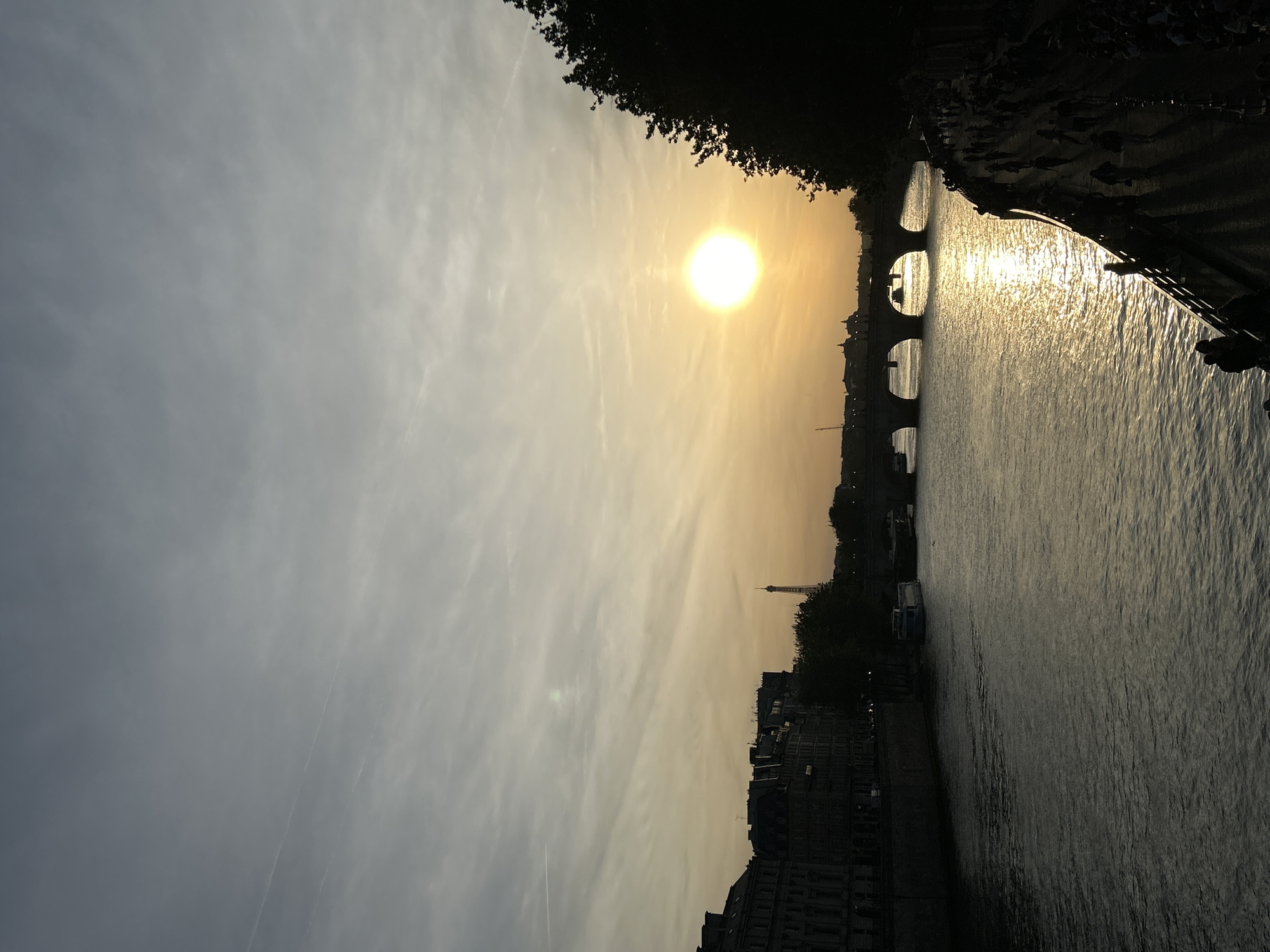 The image captures a picturesque sunset or sunrise over the Seine River in Paris, France. The sky dominates the upper portion, featuring a gradient from bright, warm yellow and orange hues near the sun on the right to cooler, muted grey and blue tones towards the left. The sun is a brilliant, circular orb, casting a strong glare on the river's surface, creating a shimmering path of light. Wispy clouds streak across the sky.
In the mid-ground, an arched bridge, characteristic of Parisian architecture, is visible in silhouette, spanning the river. Further in the distance, the iconic silhouette of the Eiffel Tower can be distinctly seen, subtly peeking above the city's skyline on the left bank. The river itself appears dark and reflective, with gentle ripples visible on its surface, particularly where the sunlight hits it.
Along the riverbanks, buildings are silhouetted against the bright sky, showcasing the dense urban landscape. Dark, leafy foliage or trees frame the upper right corner of the image, adding to the foreground elements. Small, indistinct silhouettes of what appear to be people can be faintly discerned on the riverbanks or walkways near the bridge, suggesting a public space.
The overall atmosphere is one of serene beauty, with the warm light of the setting or rising sun bathing the scene. There is no visible text in the image.