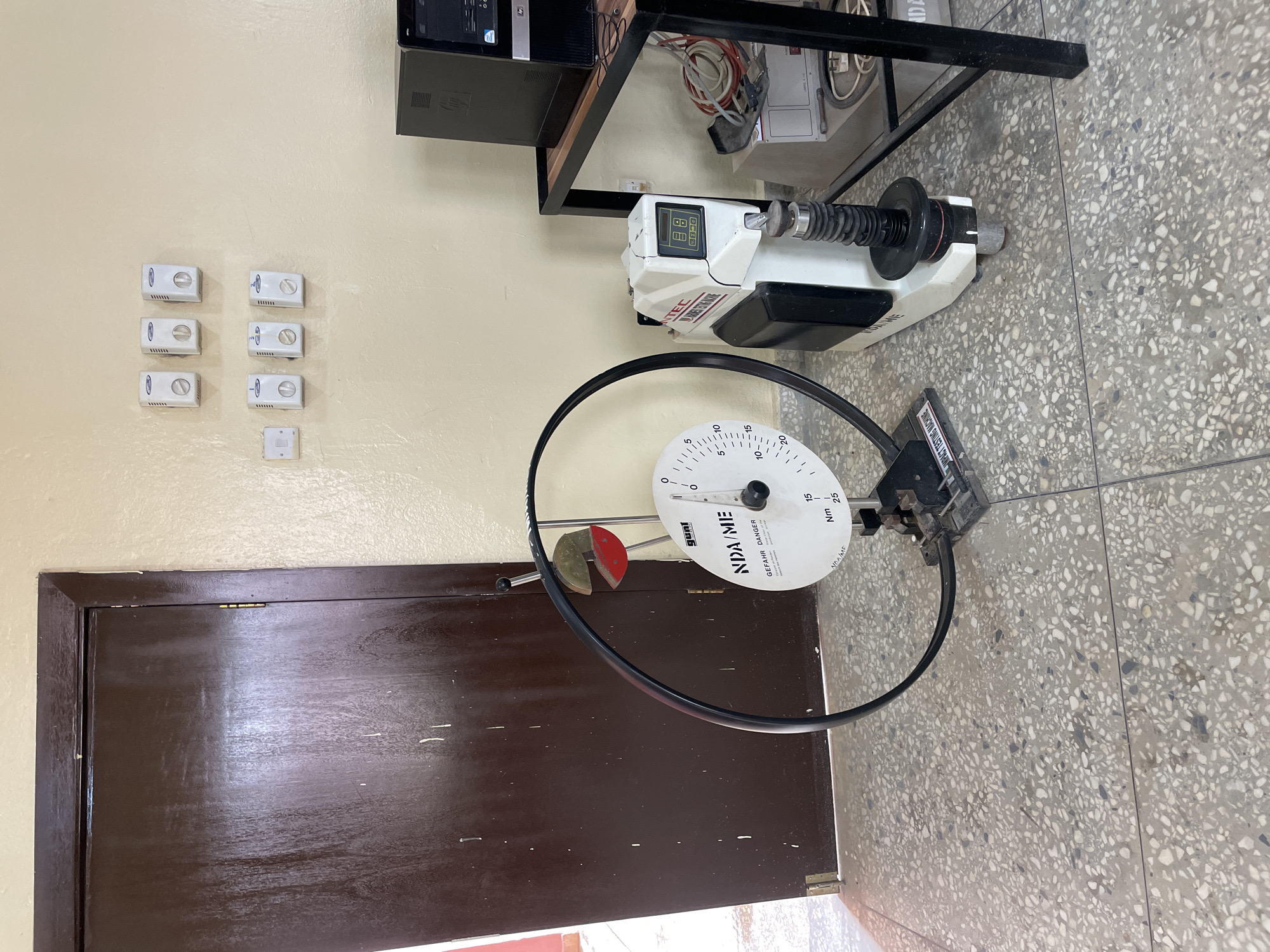 The image shows laboratory equipment in what appears to be an indoor setting. The equipment includes an impact testing machine with a circular scale labeled "NDA/ME," and another device with a digital display showing "099." The environment includes a cream-colored wall with multiple small white boxes mounted on it, a dark brown door, and a speckled gray floor. The scene likely depicts a materials testing laboratory. The location, Afaka, Nigeria, is based on information from the user query.
