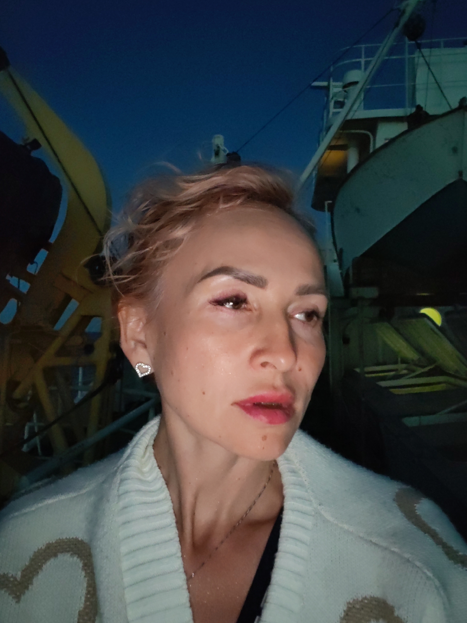 A selfie is taken in low-light conditions by a white woman with blonde hair and pink lipstick. She is wearing a white cardigan with heart designs and a delicate silver necklace. Her right ear is visible, adorned with a heart-shaped earring. In the background, there is a dark blue sky, and the structures of what appear to be ships or industrial equipment are faintly visible, illuminated by yellow light. The image is likely taken at dusk or night. The image metadata indicates it was taken in Shal'skii, Russia.