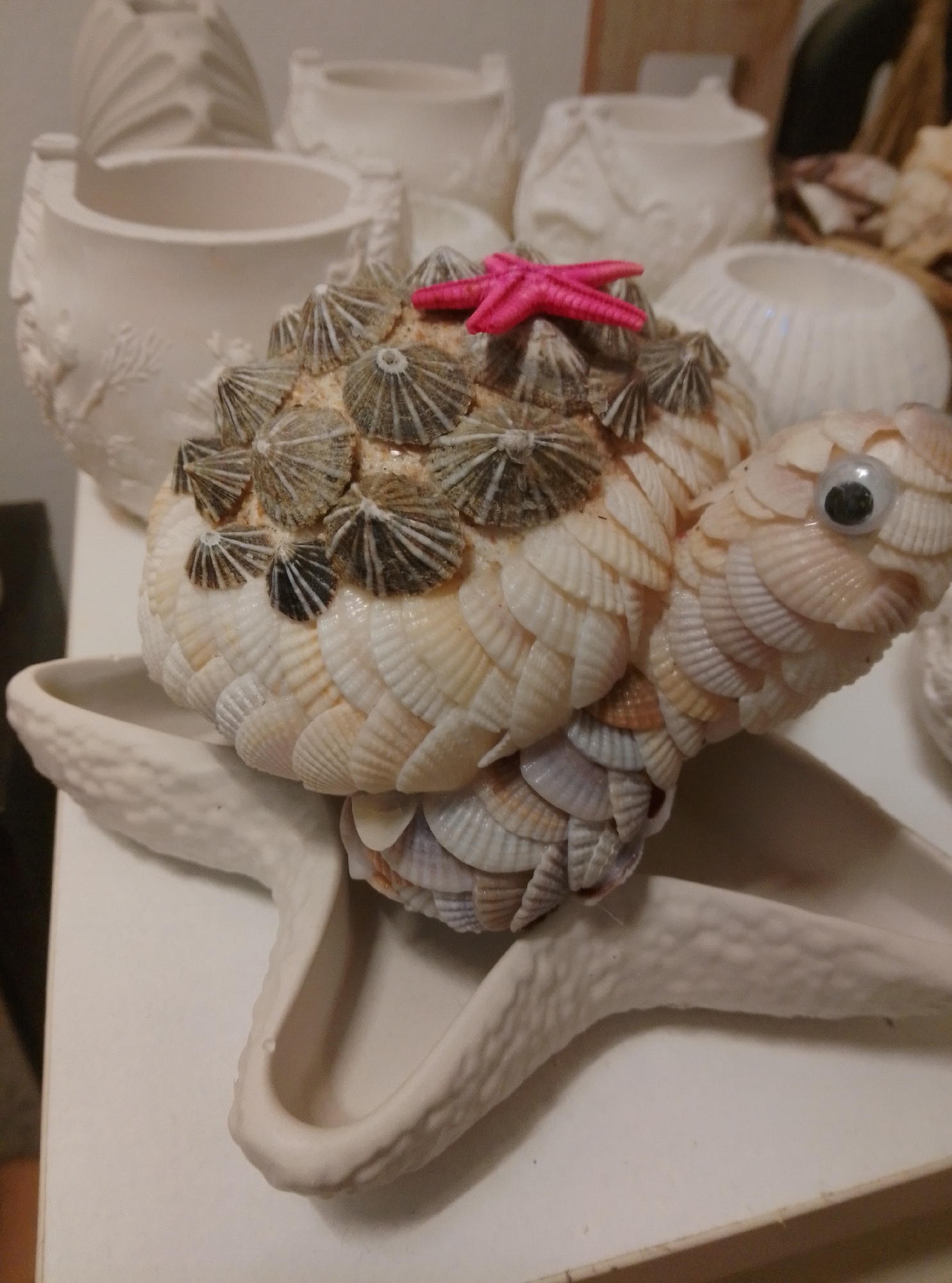 A decorative turtle sculpture, crafted from seashells and perched on a starfish base, is presented. The turtle's shell is adorned with numerous small, conical shells, likely barnacle clusters, and a small pink starfish embellishment is affixed to its upper shell. The turtle has a single googly eye and is composed of overlapping, small white and beige seashells, resembling scales. The starfish base is a creamy white ceramic with a textured surface. In the background, several white ceramic pots or vases are visible, some with decorative relief patterns. The scene appears to be indoors, possibly in a home or workshop, with a neutral-colored surface beneath the objects. The image does not contain information about the city of Wedel, Germany.