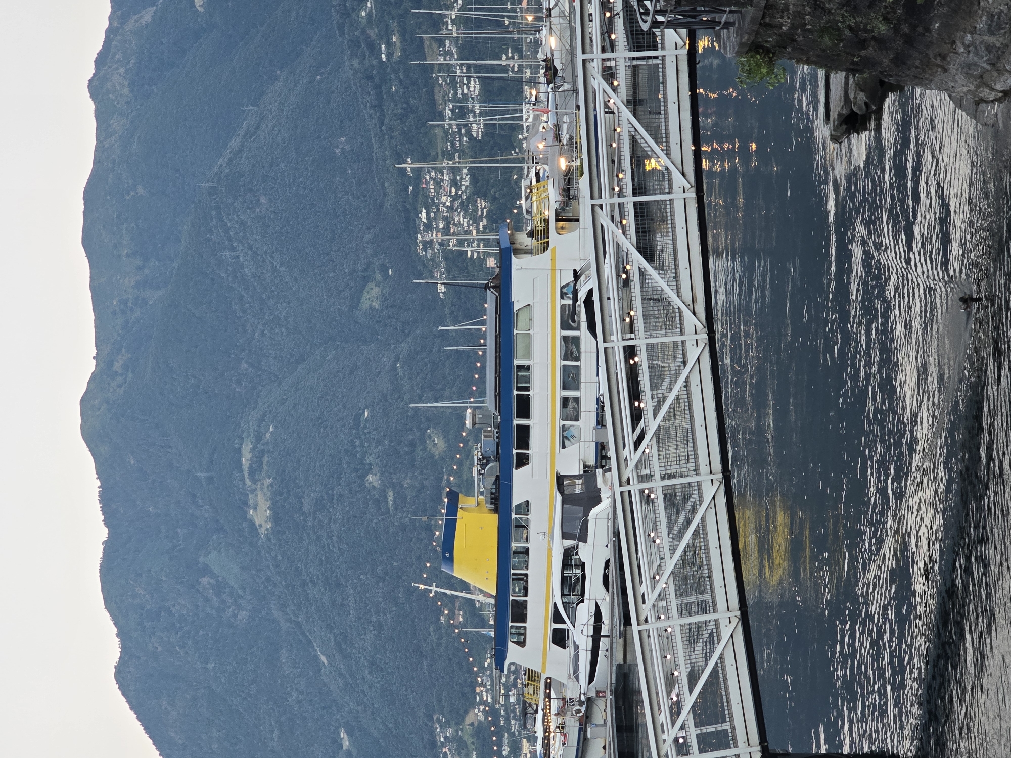 A large white and blue passenger ferry with a yellow funnel is docked at a pier in Locarno, Switzerland. The ferry is illuminated by strings of lights. In the foreground, a white pedestrian bridge spans the water. The background features a large, tree-covered mountain and a town built on its slopes. The water is dark and reflects the lights from the bridge and ferry. A duck is swimming in the water in the lower left corner.