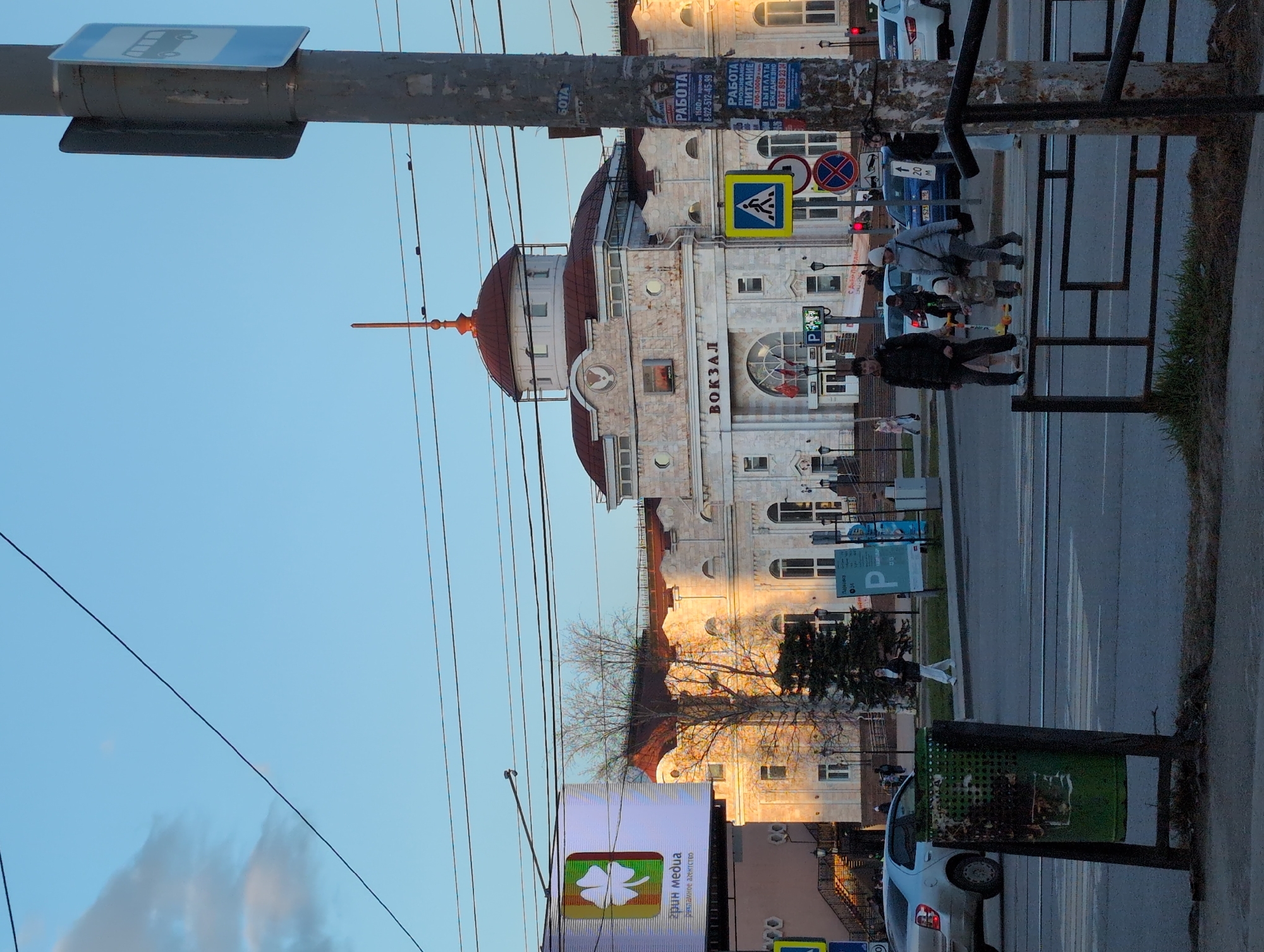 The image depicts the Izhevsk Railway Station building in Izhevsk, Russia. The station is a multi-story structure with a distinctive red dome. In front of the building is a street with pedestrian crossings. People are walking across the street, including a family with a child. The surrounding environment includes utility poles with wires, signage, and buildings in the background. A bus stop sign is visible on a pole. A green advertisement sign with the text "грин медиа" is visible on the lower left. Cars are parked along the roadside.