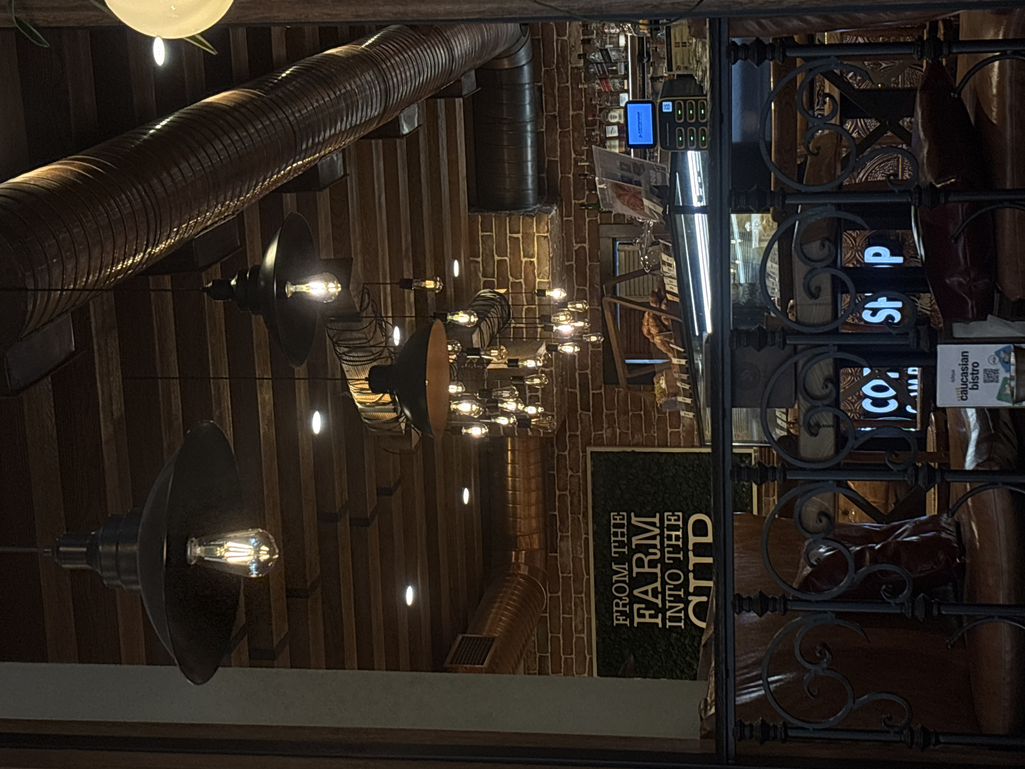 The image captures an interior view, likely of a restaurant or café in Yerevan, Armenia, as indicated by signage mentioning "Caucasus bistro." The scene is dominated by industrial-style elements: exposed ceiling beams, a large ventilation duct, and pendant lights with Edison bulbs. The lights illuminate the space, creating a warm ambiance. Through a decorative wrought-iron divider, one can glimpse seating areas with leather-upholstered furniture. A sign reading "FROM THE FARM INTO THE CITY" suggests a focus on locally sourced ingredients. A cash register and some merchandise are visible in the background.