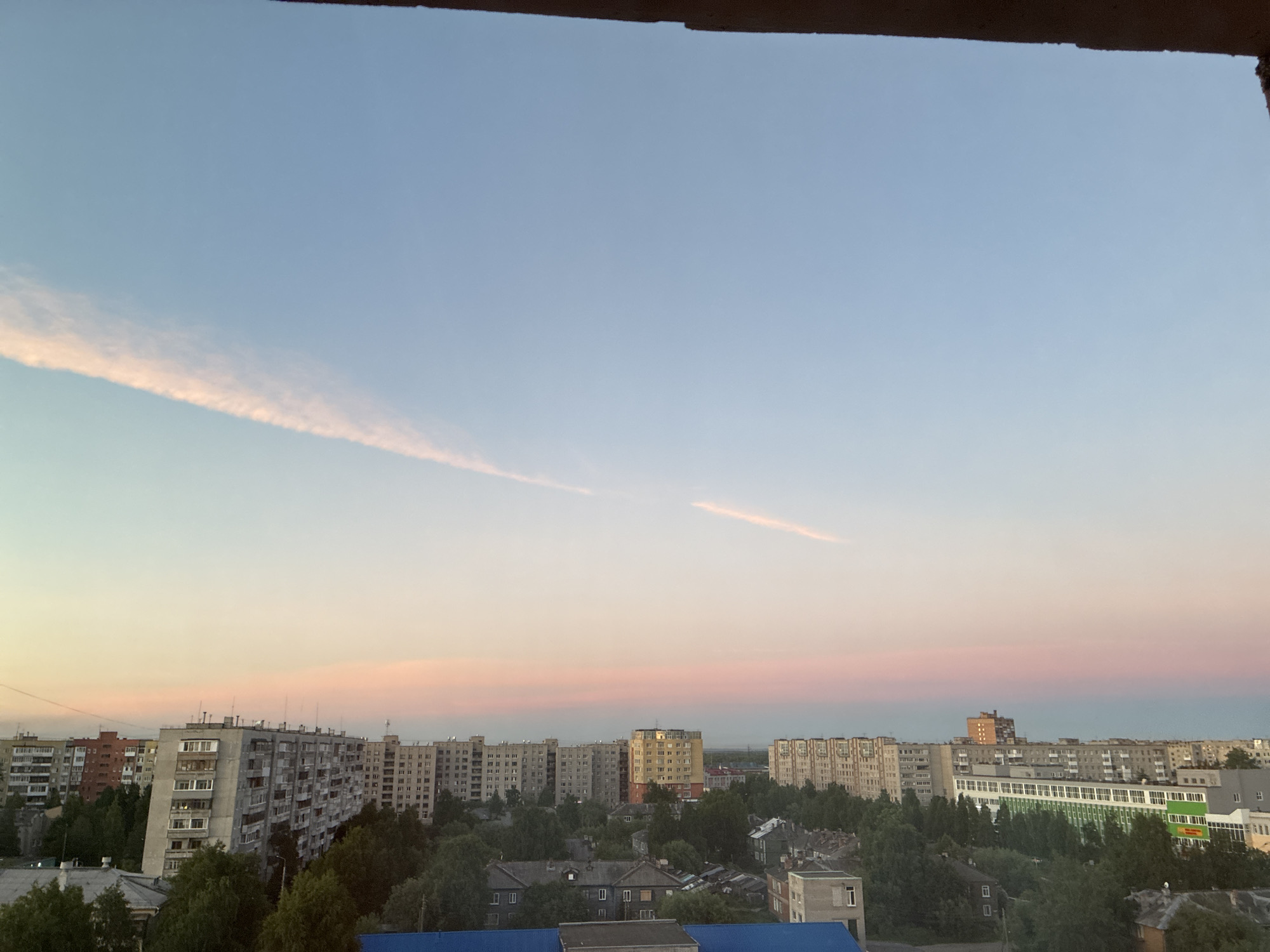 An elevated, wide-angle, eye-level shot captures a cityscape during what appears to be twilight or dawn. The sky is a gradient of soft blues and pinks, with a faint, elongated contrail streaking across the upper portion of the frame. Below the sky, a dense arrangement of apartment buildings and smaller houses occupies the midground and foreground. The architecture is predominantly Soviet-era concrete blocks, varying in height and color, with numerous windows visible. Dense green trees are interspersed between the buildings, suggesting a city with significant green spaces. The overall impression is one of a residential urban environment bathed in the gentle light of the transitioning sky.