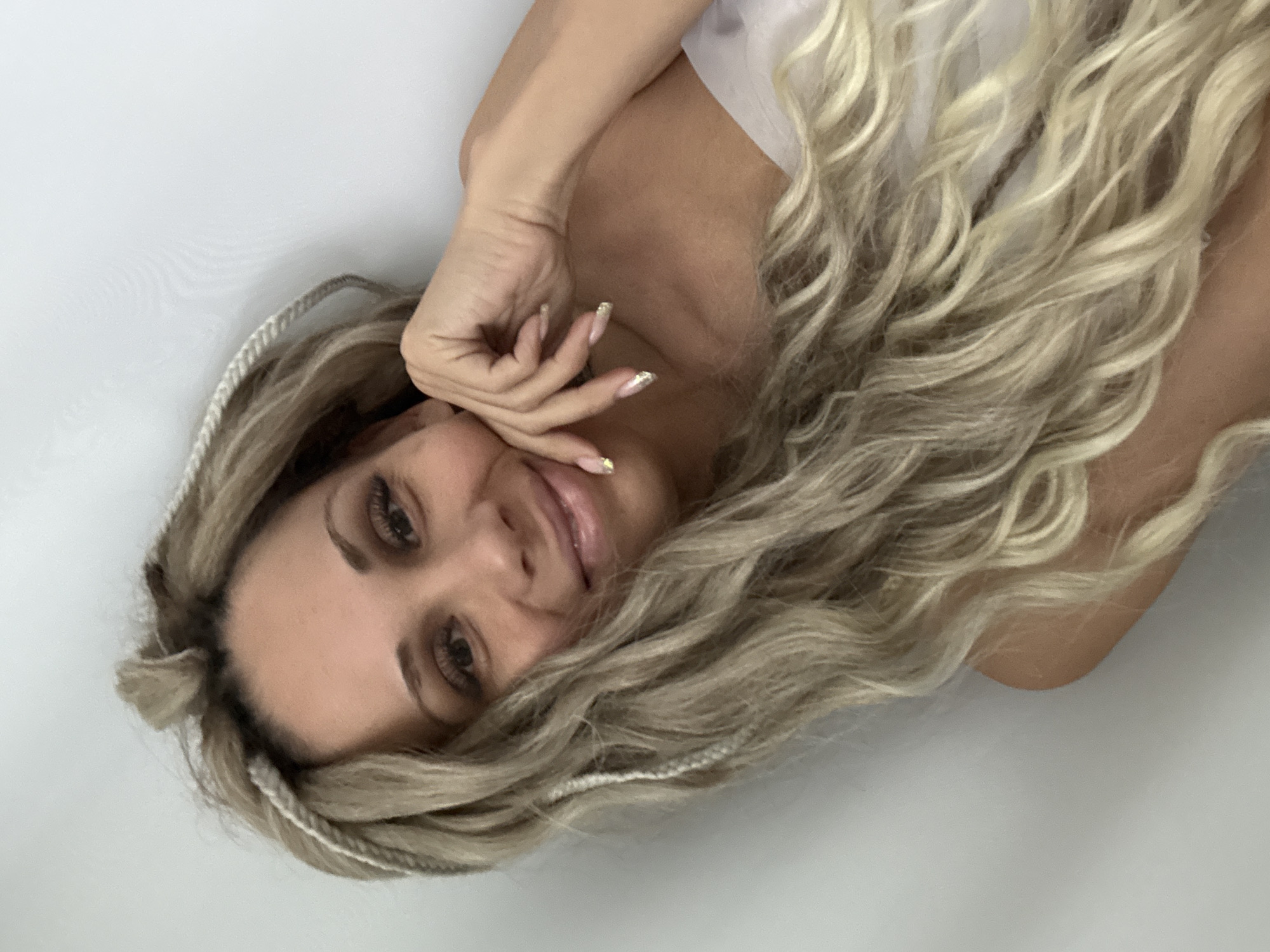 A woman with long, wavy, blonde hair and partial white braids lies with her head tilted on a white surface. She has her hand near her face, displaying manicured nails with gold accents. The image suggests an indoor setting, but there are no other environmental details to infer a specific location like Rusanovo, Russia.