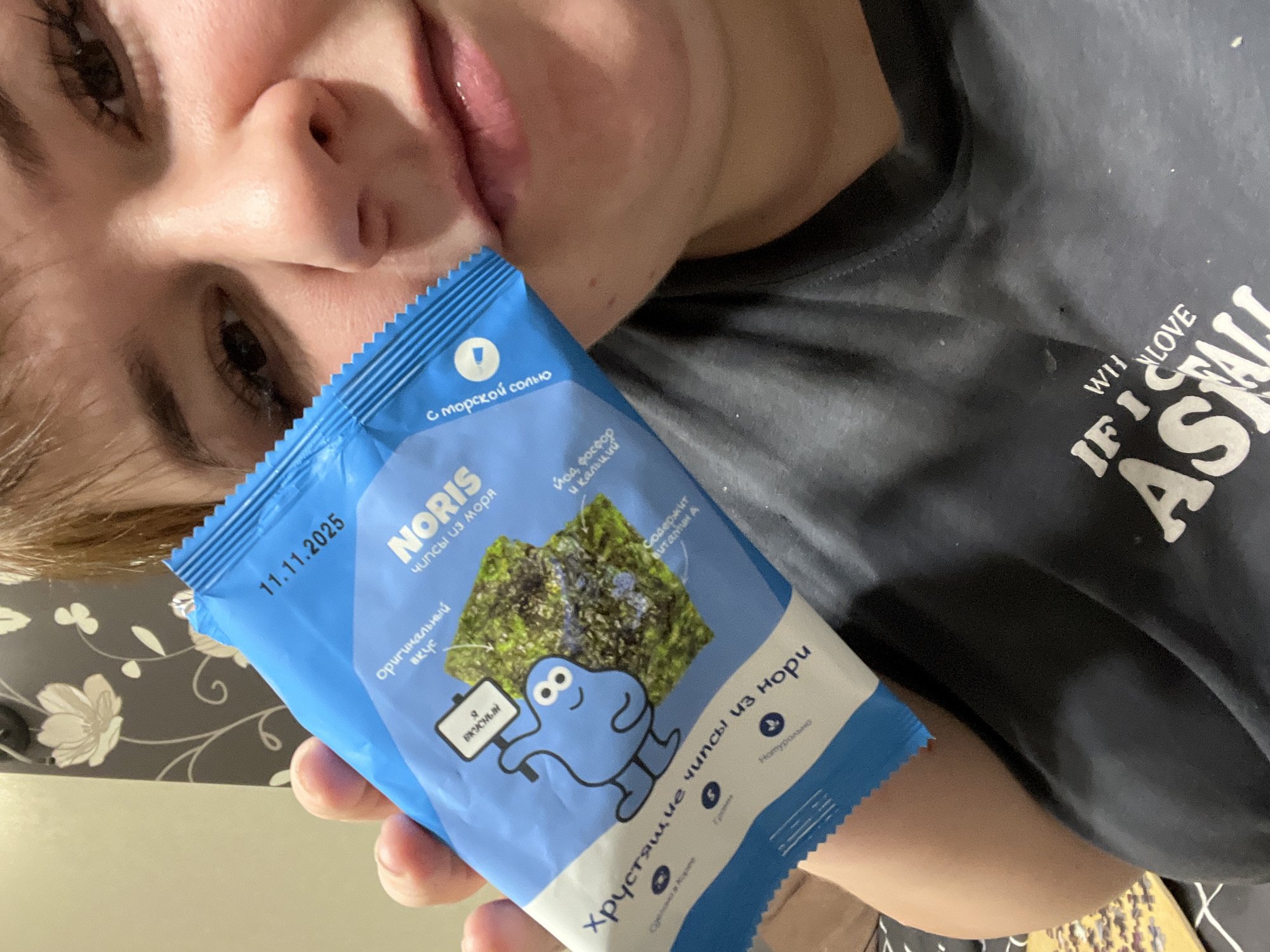A close-up indoor photograph depicts a person, primarily showing their upper face (eyes, nose, and mouth) and upper torso, holding a blue and white flexible pouch of "NORIS" brand nori chips. The person, likely female, has light brown hair and is wearing a dark grey t-shirt with partially visible white text reading "IF I WILL LOVE ASALL". The food package, held by the person's right hand, prominently displays an expiration date of "11.11.2025" and features a cartoon blue character, an image of dried nori, and Cyrillic text identifying it as "Crispy Nori Chips" with "Original Taste" and "Sea Salt." The background reveals an interior residential setting with dark grey wallpaper featuring a white floral pattern behind the person's head. In the lower left, a surface with a black and white geometric pattern holds a small pile of loose puzzle pieces. No specific location within the city of Chaikovsky, Russia, is discernible beyond it being an indoor residential environment.