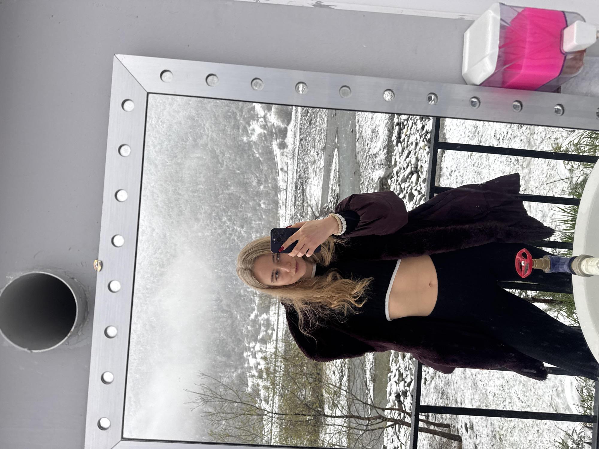 A young woman is taking a selfie in a mirror. The mirror has a metal frame with circular cutouts along the edges. The reflection shows a snowy landscape with trees and a river or stream. The woman is wearing a dark top and a fur-like coat. A water valve and container of tissues are visible near the mirror. The location is identified as the city of Unknown, Unknown.