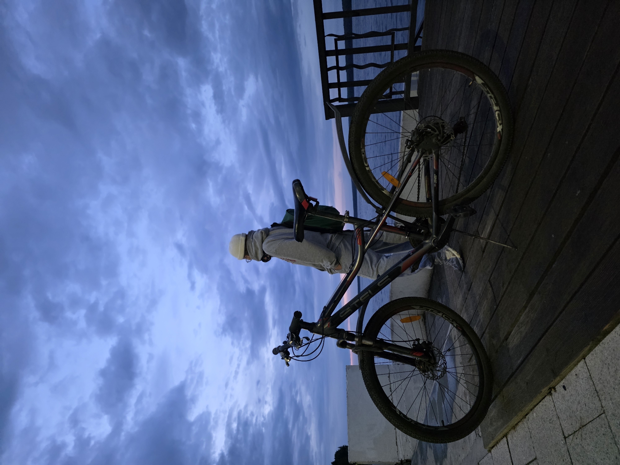A person and a bicycle are next to a railing with a body of water in the background. The person is wearing a light-colored beanie and a gray sweatshirt with matching sweatpants. The bicycle is black with red accents and is leaning against the railing. The sky is filled with gray and blue clouds. The location may be the city of Chaikovsky, Russia.