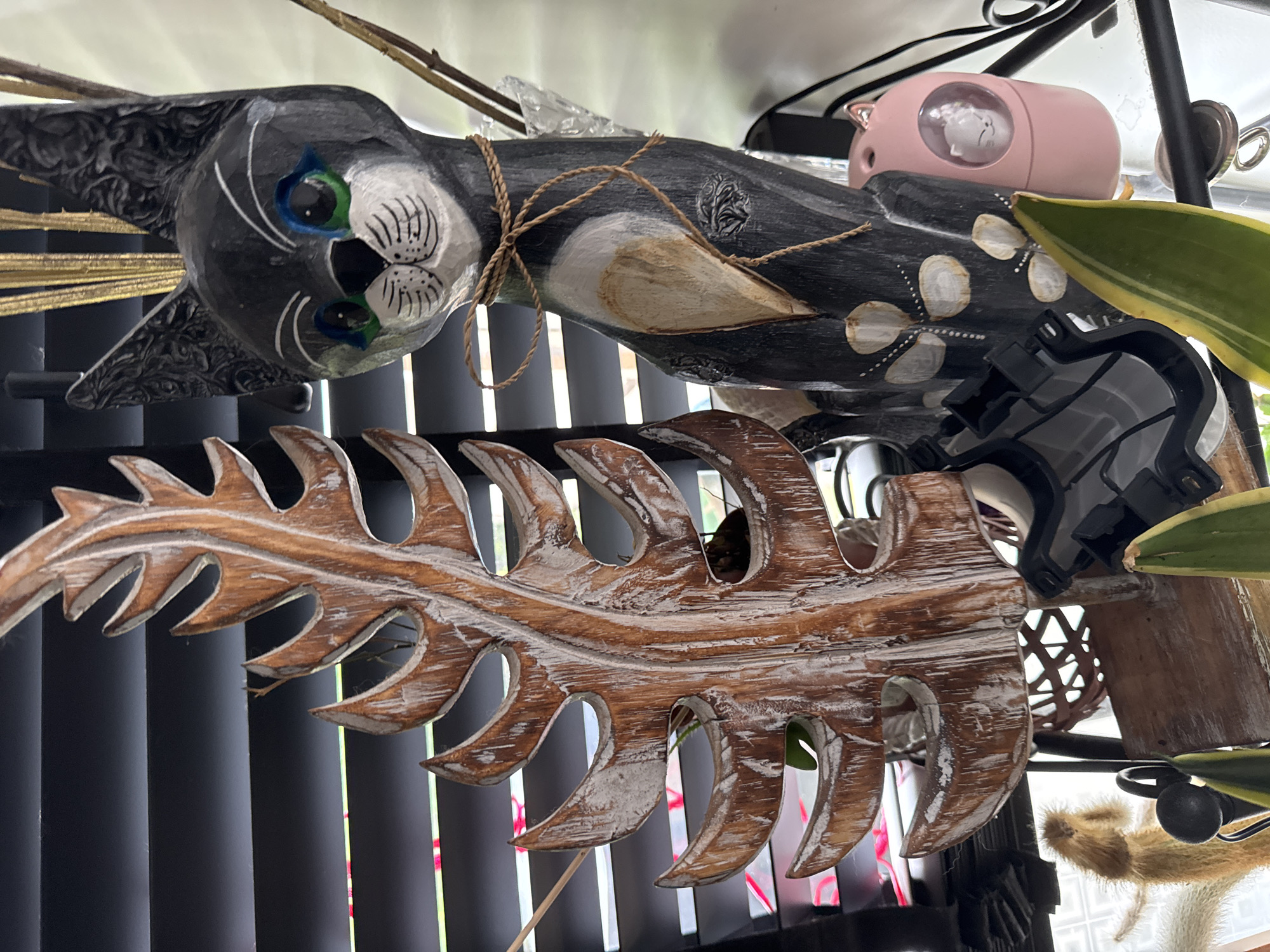 The image depicts an indoor static display featuring two primary decorative wooden sculptures. On the left is a tall, vertical carved wooden sculpture shaped like a serrated tropical leaf, exhibiting a distressed finish with brown wood tones and significant white paint or whitewash. To the right, a slender, dark grey or black painted wooden cat figurine stands upright. The cat features green eyes with blue eyelids, white whiskers, a black nose, a white muzzle and chin, and white circular patterns on its lower body. Intricate carved patterns resembling floral or swirl motifs are present on its ears and parts of its body. A thin, light brown twine is tied in a bow around the cat's upper chest. Behind the sculptures, dark grey horizontal window blinds are partially visible, with faint glimpses of bright green and pink/red light or objects showing through the slats. In the lower right foreground, a small pink cat-shaped object with a clear circular window displays a small white cat figurine inside. Parts of green plant leaves, one with yellow variegation, are visible in the lower foreground. A dark, angular object with geometric shapes is positioned below the wooden leaf sculpture, and a portion of a brown woven basket is visible in the lower-middle background. Gold-colored string-like decorative elements extend upward from behind the wooden cat's head. The scene is devoid of human presence or active interaction.