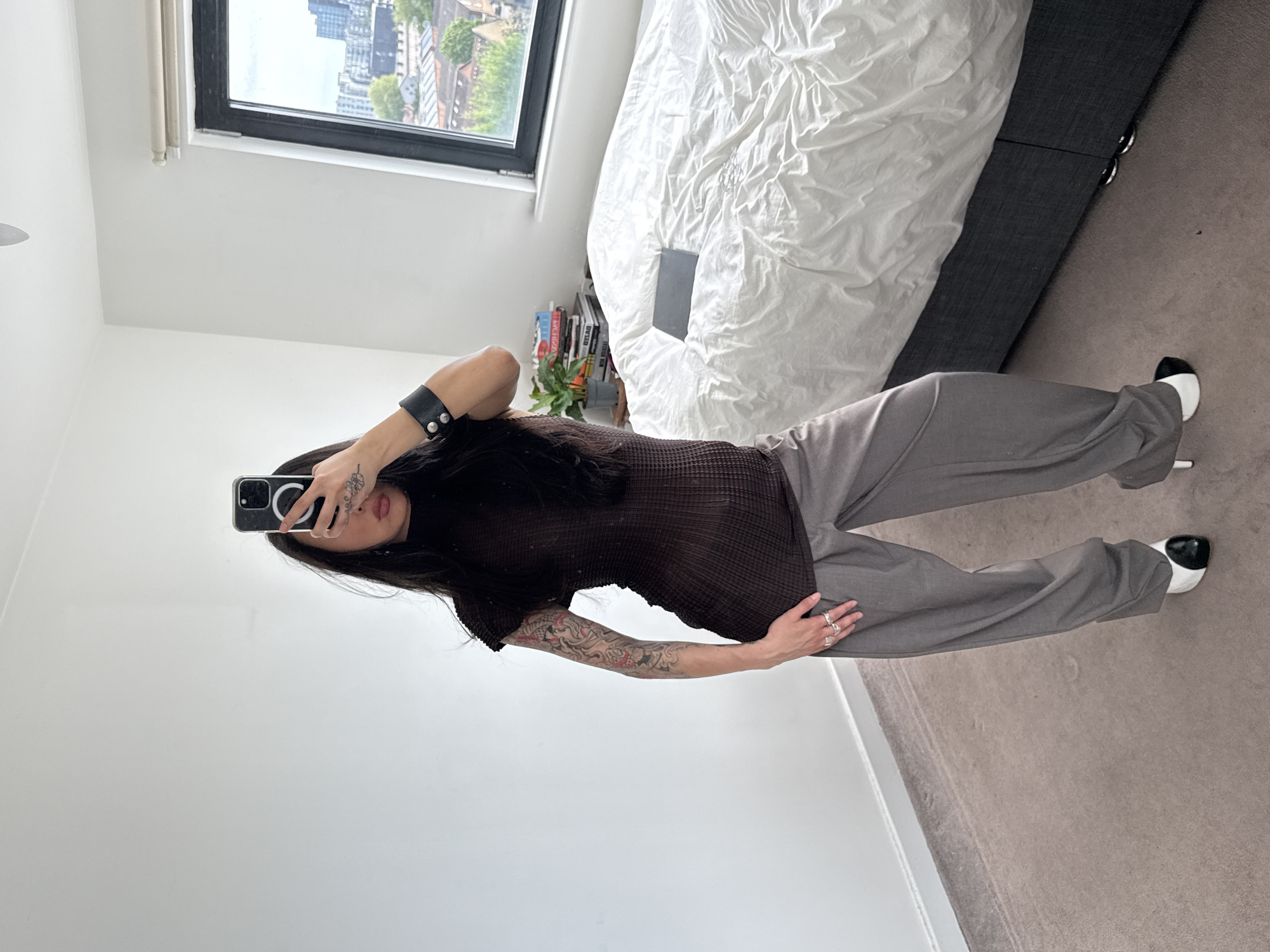 The image displays a mirror selfie taken by a person in what appears to be a modern bedroom or studio apartment. The image is oriented 90 degrees clockwise.
The central subject is a person, likely female, with long dark hair, holding a black smartphone with a metallic ring attachment to capture their reflection. Their face is partially obscured by the phone, but their mouth and chin are visible. They are wearing a dark brown, sheer, ribbed or pleated short-sleeved top, paired with light grey wide-leg trousers and distinct white and black two-tone high-heeled shoes. Notable details include a prominent sleeve tattoo on their left arm, featuring koi fish and other intricate patterns, as well as smaller tattoos on their left hand and fingers, adorned with multiple rings. A black wristband with two silver studs is visible on their right wrist.
The setting is an indoor space characterized by minimalist white walls and a light brown/grey carpeted floor. In the background, a large bed with a white, wrinkled duvet cover and a dark grey upholstered frame is visible. A dark rectangular object, possibly a book or tablet, rests on the duvet. To the left, a large window with a black frame offers a view of a cityscape, featuring modern buildings and some greenery, indicative of an urban environment like London, United Kingdom. Below the window, a stack of books is visible on a surface, with the partial word "GUCC" discernible on one spine, alongside a small green plant.
The activity depicted is the act of taking a mirror selfie. The time of day appears to be daytime, with natural light filtering through the window, suggesting an overcast or partially cloudy sky. The person's expression is not fully visible due to the phone's position.