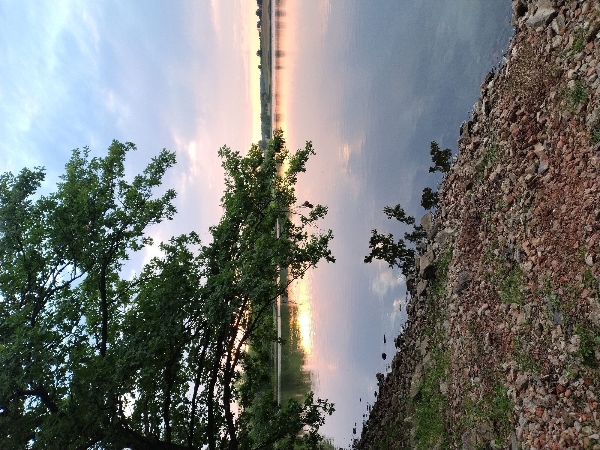 This is a photograph of a lake in Kestřany, Czechia, at dusk. The sky is a pale blue with soft pink and orange hues reflecting on the water. Lush green tree branches overhang the left side of the frame, casting shadows on the lake. The shore in the foreground is rocky and uneven, with scattered green vegetation. The water is calm and reflective, showing a mirror image of the sky and the surrounding trees. In the distance, the far shore of the lake is lined with trees and rolling hills, suggesting a rural or park-like setting. There are no people or animals visible in the scene.