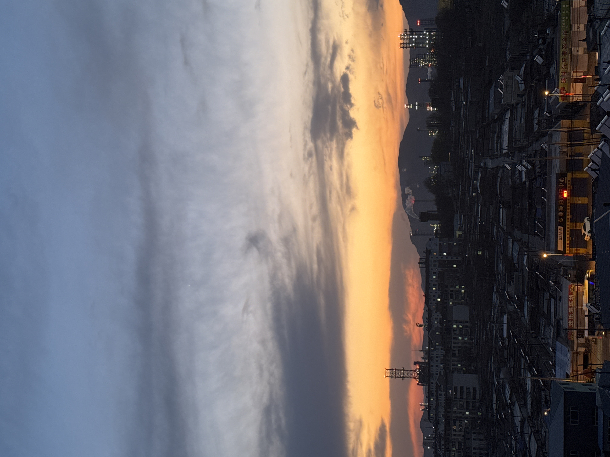 The image captures a view of Zhongtun, China at dusk. On the right side of the frame, a densely populated urban landscape is visible, with numerous buildings illuminated by streetlights and windows. The architecture appears utilitarian, likely residential or commercial structures typical of a bustling Chinese city. The left side of the frame showcases the sky, with a gradation of colors transitioning from a dark blue-gray to a warm orange-yellow near the horizon. The sky is partially covered with clouds. The interplay of the natural twilight sky and the artificial lights of the city creates a distinct visual contrast.