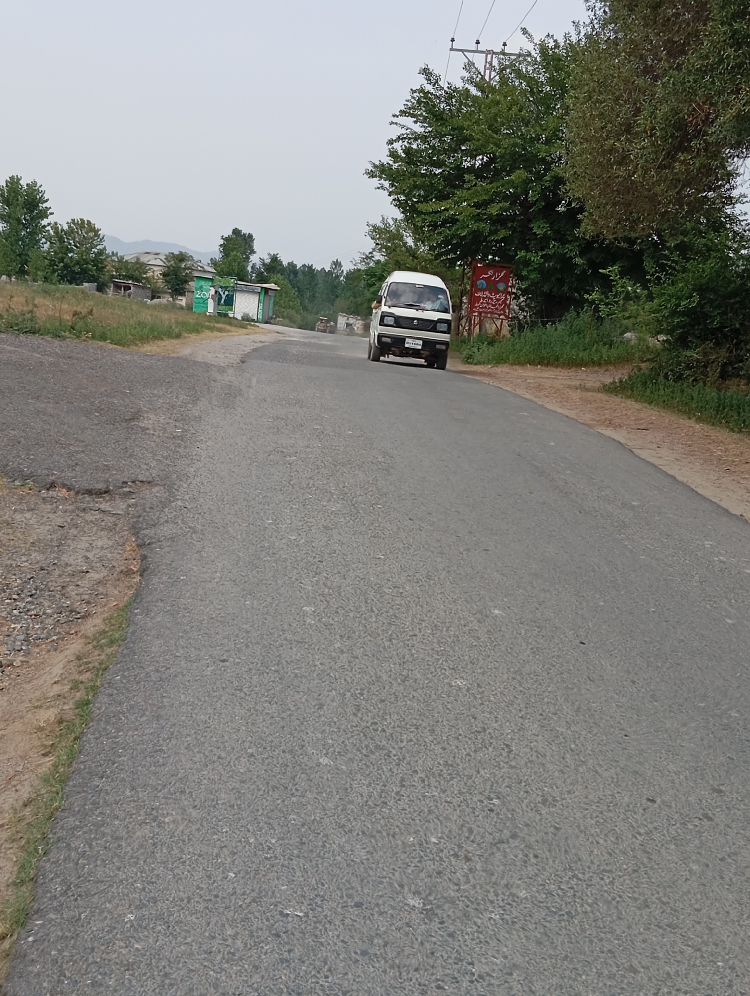 A white minivan with a license plate reading "G11-1664" drives on a paved road. The minivan is in the center-right of the image and is traveling away from the viewer. To the left of the road is a dirt shoulder with some grass growing on it. Further to the left is a grassy hill with some buildings in the background. To the right of the road is more grass and trees. In the far background, mountains are visible. The sky is overcast.