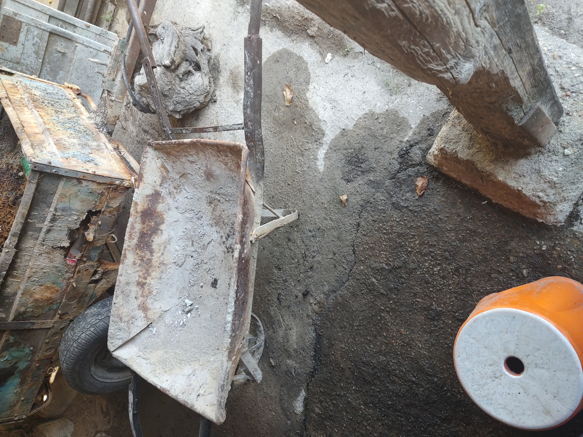 An overhead, slightly angled view of a weathered, dusty, and rusty scene in Kochmar, Bulgaria, depicts construction or demolition debris. In the foreground, a discarded orange and white plastic stool sits on a dark, wet, and cracked concrete surface. To the right, a thick, rough-hewn wooden post leans into the frame, supported by a stone base. The background features a derelict, heavily rusted metal cart, possibly a trailer, with a single tire visible. A wheelbarrow, filled with light-colored dust and debris, is positioned in front of the cart. The ground is a mixture of dirt, gravel, and concrete, with scattered leaves and debris. The overall impression is one of neglect and disuse, typical of a rural or industrial storage area.