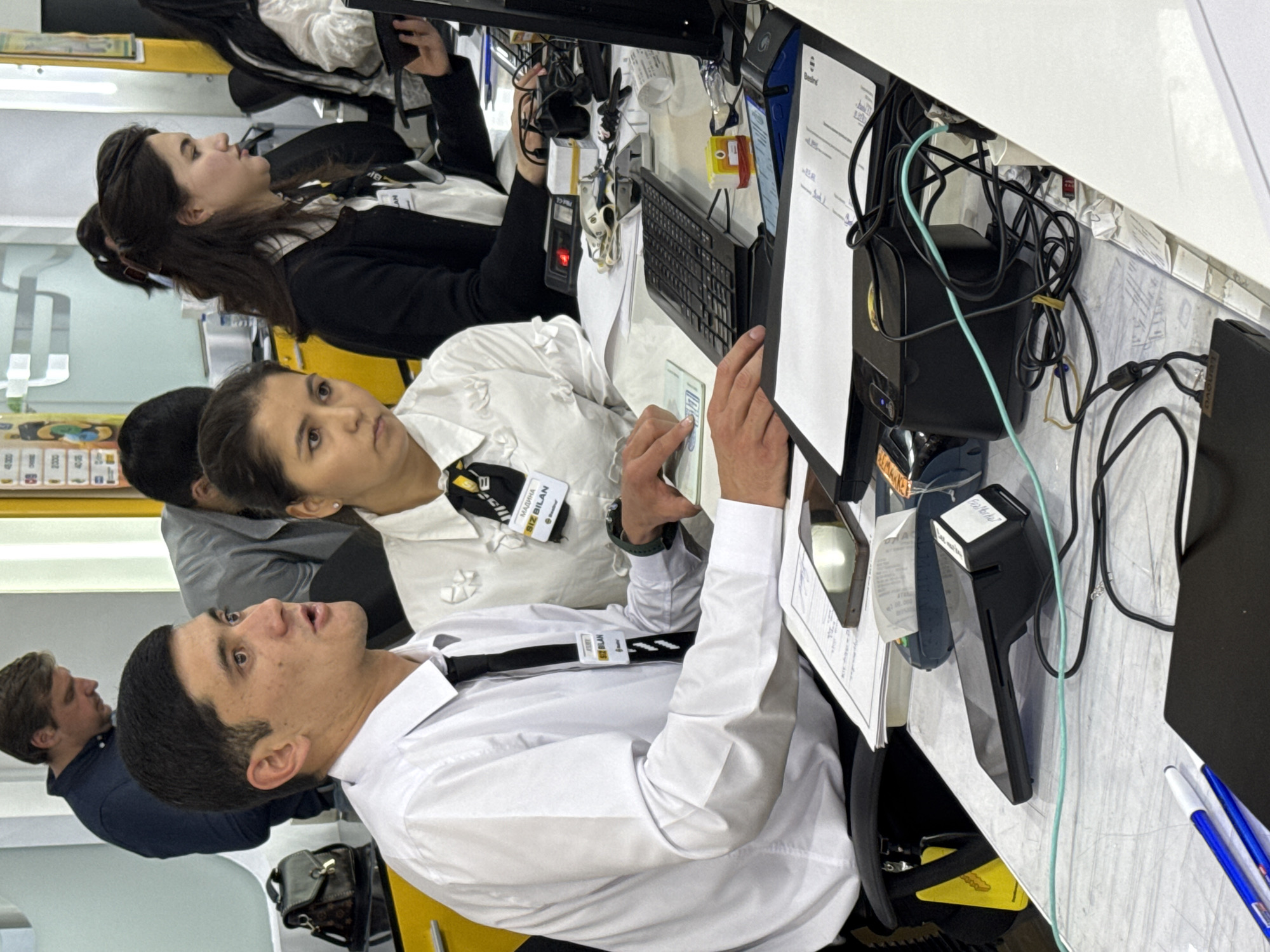 Inside what appears to be an office environment in Tashkent, Uzbekistan, several individuals are situated at desks with computer monitors. The scene features three people prominently: a man wearing a white shirt and black tie, and a woman in a white blouse. They both have name tags written in Uzbek that include the name "Siz Bilan," suggesting they are employees. The man is looking up in surprise. Both appear to be engaged with some form of paperwork, potentially passports, based on the documents visible near their hands. They sit alongside another woman in a black sweater who is looking to the left.
Additional objects include computer hardware (monitors, keyboards), a card reader, a printer, wires, pens, and a stapler, all indicative of a functioning office workspace. The desk is cluttered with paperwork and office supplies. The scene suggests a service-oriented business that deals with official documentation.
