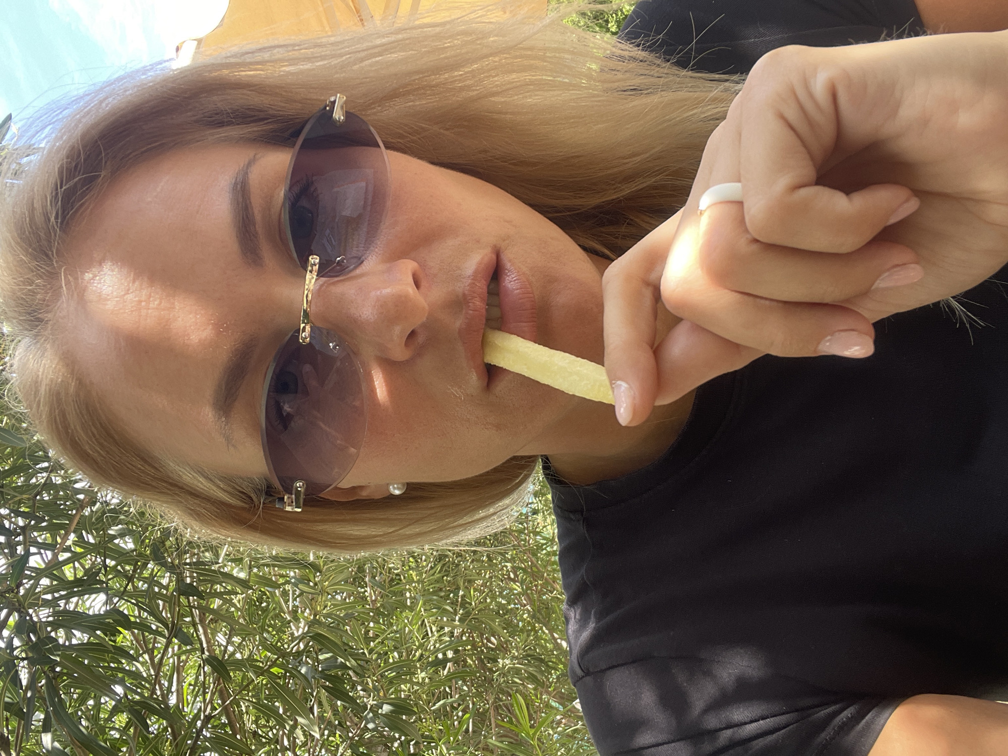 A young woman is shown in a close-up shot, possibly a selfie. She has blonde hair and is wearing sunglasses and a black shirt. She is holding and eating a french fry. In the background, there is greenery suggesting an outdoor setting, potentially a park or garden. The city location is Sochi, Russia.