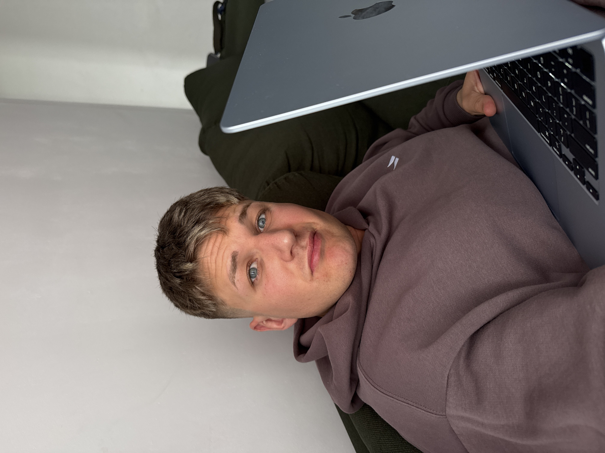 A young, fair-skinned man with short blonde hair and blue eyes is lying on a dark green cushioned surface, possibly a sofa or bed. He is wearing a light brown hoodie. He is holding a silver laptop, with the Apple logo visible on the lid. The laptop keyboard is also visible. The background is a plain white wall. The setting does not obviously indicate Plovdiv, Bulgaria; the scene is indoors and could be anywhere.