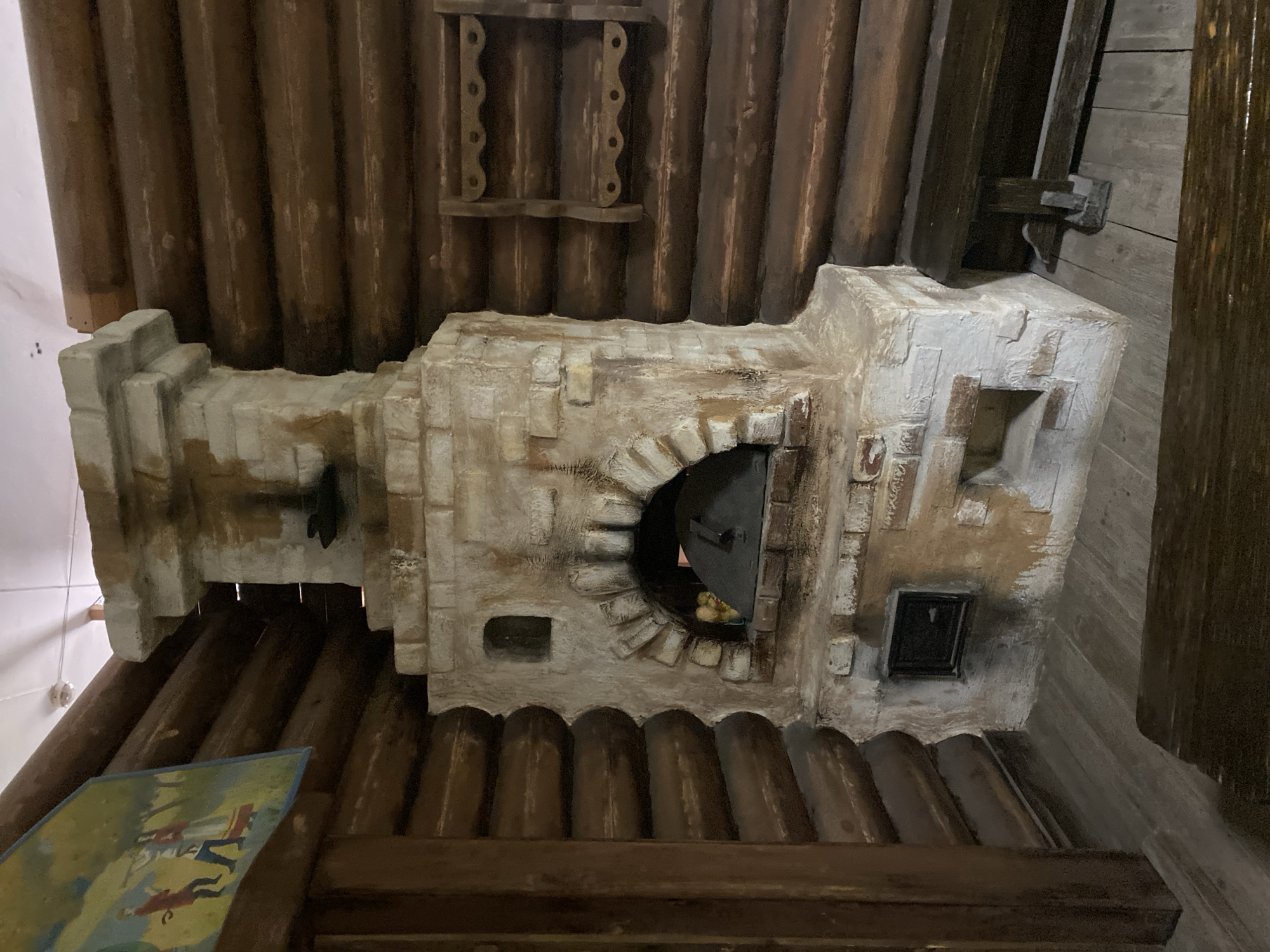 The image shows a detailed model of a traditional Russian stove, possibly located in a historical or cultural context, likely at a museum or historical site in Yekaterinburg, Russia. The stove is constructed of what appears to be painted brick or stone and features an oven door ajar revealing what look like onions inside, an ash pit access door, and what could be a rotating damper. The background features log cabin style walls and ceiling, along with other small decorative wood features. A partially visible picture or sign is on the ground in front of the stove. The lighting is ambient, with some shadows indicating a diffused light source.