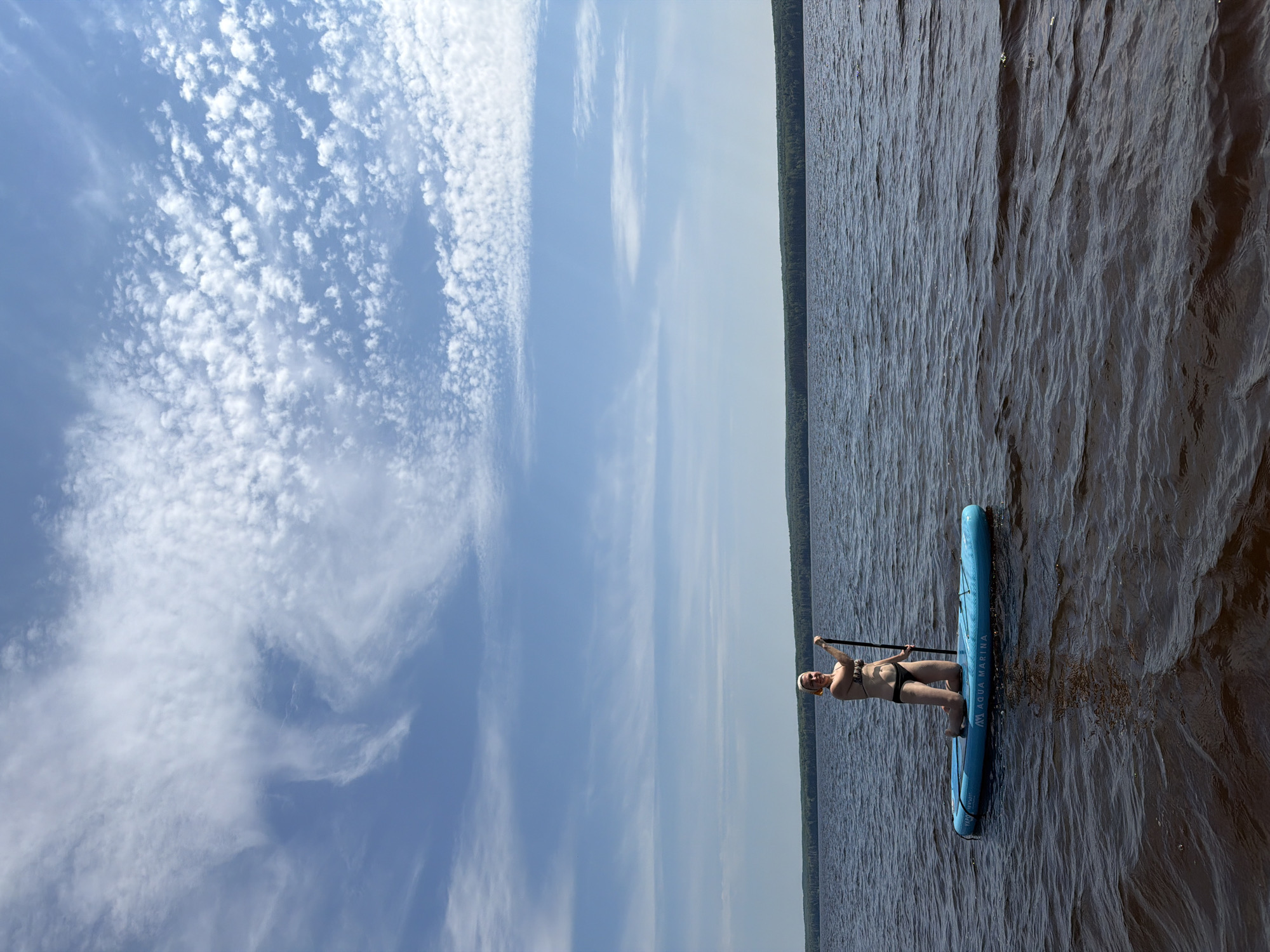 A woman in a bikini kneels on a blue stand-up paddleboard in dark water. The paddleboard has the brand name "Aqua Marina" visible. The woman is holding a black paddle. She is smiling and looking towards the camera. The background shows a vast body of water, a distant tree line, and a sky filled with scattered white clouds. The water is dark brown with some ripples. The scene appears to be a natural outdoor setting, possibly a lake or large river, under a bright sky. The location is specified as Chaikovsky, Russia.