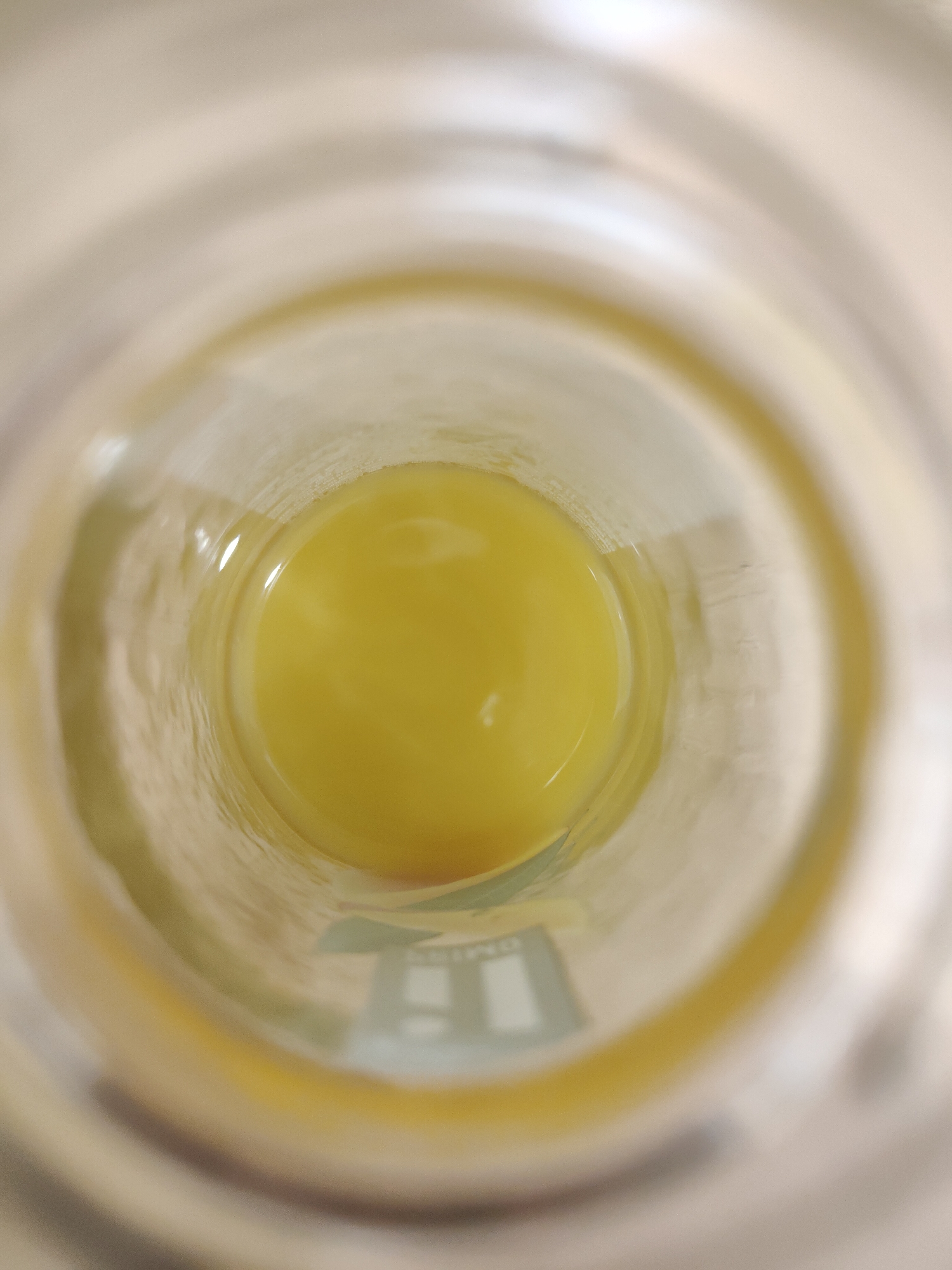A top-down view inside a cylindrical glass containing a viscous, opaque yellow liquid. A reflection of an indeterminate object is visible at the bottom of the glass. The location is Unknown, Unknown.