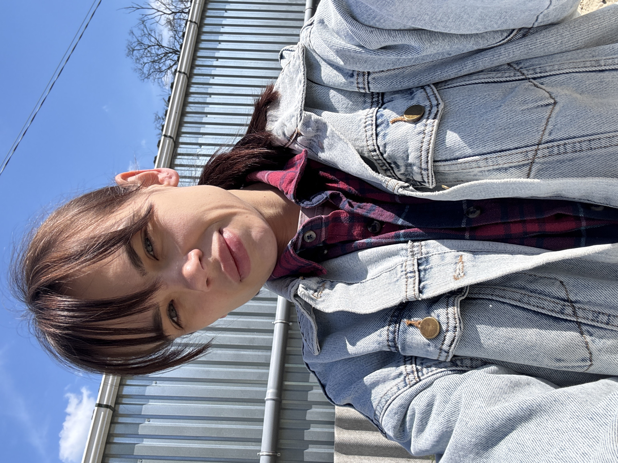 A young woman is posing for a selfie in what appears to be an outdoor environment. She has brown hair with bangs and is wearing a denim jacket over a plaid shirt. In the background, there's a corrugated metal fence or wall, a bright blue sky, and tree branches without leaves, suggesting it might be late fall or winter. While there is no definitive information to confirm the image's location as Rostov-on-Don, Russia, the general architectural and environmental cues are consistent with urban settings. There are no other people or subjects directly visible in the image. The main action is the woman taking a selfie.