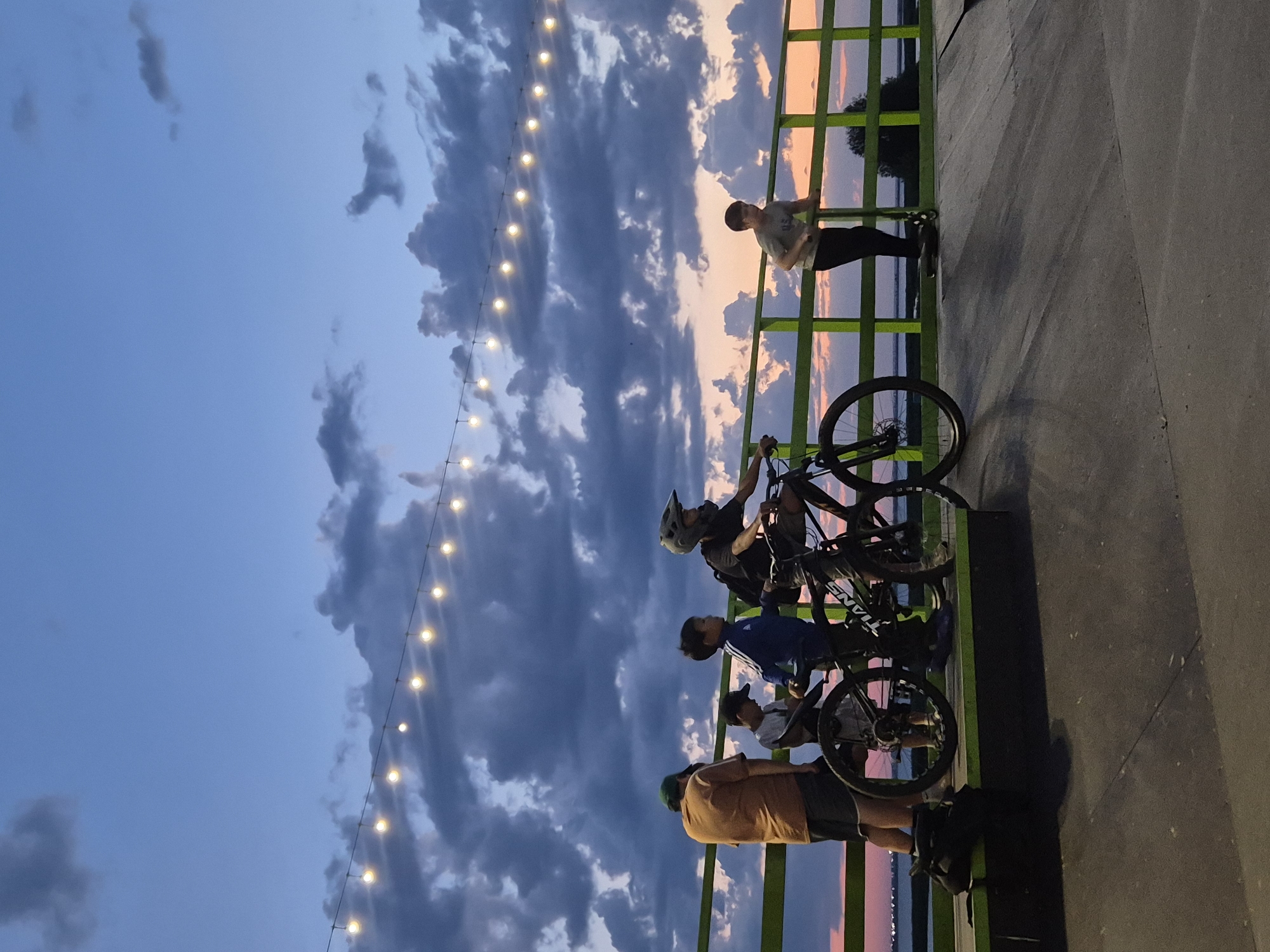 The image depicts an outdoor scene at dusk, likely in Kokshetau, Kazakhstan, given the context. A group of individuals, primarily young males, are gathered at what appears to be a skate park or a recreational area near a body of water.
In the foreground, there is a concrete ramp with a green railing. On the ramp, two boys are on bicycles. One boy, wearing a helmet, is on a black bicycle. Another boy is standing next to a bicycle, and a third boy is seen standing beside him. An adult male, wearing a brown shirt and shorts, is also present, standing near a black bag on the ground. Further to the right, a young boy is on a scooter, leaning on the green railing.
The background shows a body of water with a distant shoreline and trees. The sky is a dramatic mix of deep blues and grays, with scattered clouds illuminated by the fading light of sunset, casting hues of orange and pink. A string of lights is strung across the sky, adding a festive ambiance to the scene. The overall atmosphere is one of casual recreation and relaxation during the twilight hours.