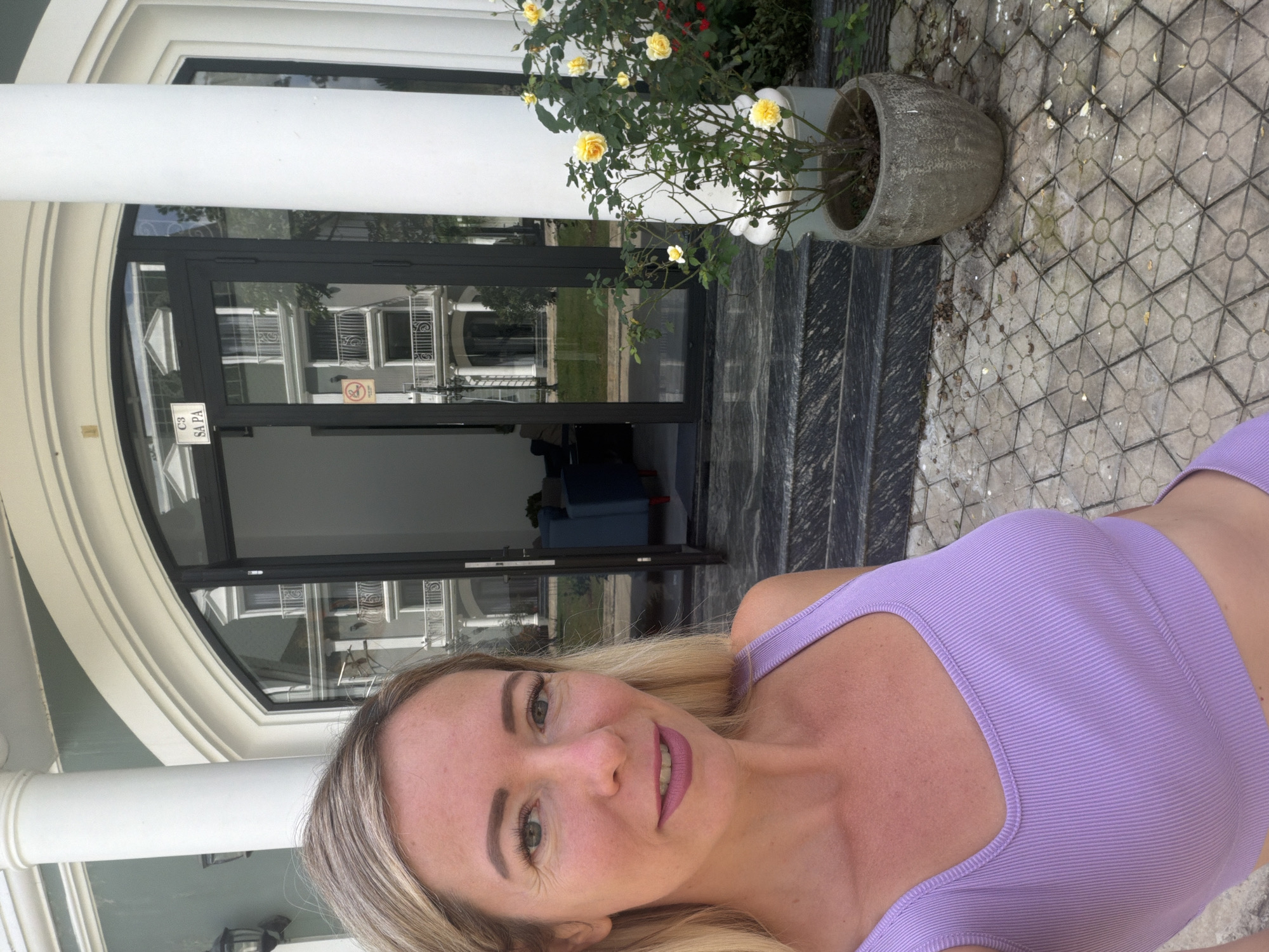 A woman with blonde hair and green eyes smiles at the camera, wearing a lavender crop top. She is standing on a patterned paved walkway in front of a building. The building has large glass doors with balconies reflected in them, and a sign that reads "C3 SAPA". To the right of the entrance, a potted rose bush with yellow flowers is visible. The walkway is bordered by a dark granite staircase. The setting appears to be an outdoor area in the city of Xuan Truong - Da Lat, Vietnam.