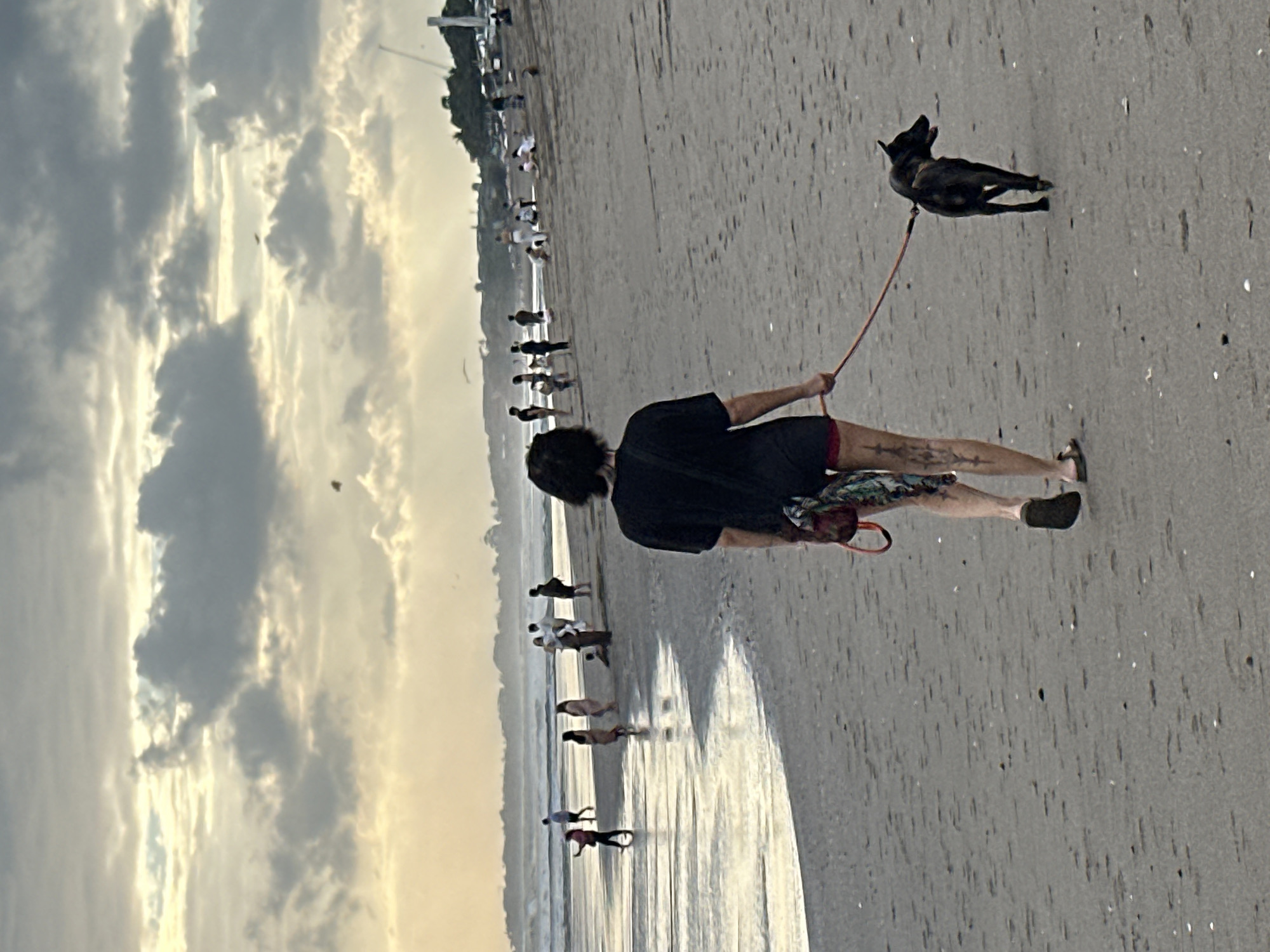 A person walks a dog on a beach in North Kuta, Indonesia, during an overcast day. The person, facing away from the camera, wears a black t-shirt and shorts, with a patterned cloth bag slung over their shoulder. Their legs appear sandy. The dog, a dark-colored breed with pointed ears, is on a leash. In the background, several people are scattered across the sand and wading in the shallow water. The ocean has moderate waves, and the sky is filled with dramatic clouds. Palm trees and structures are visible on the distant shoreline.