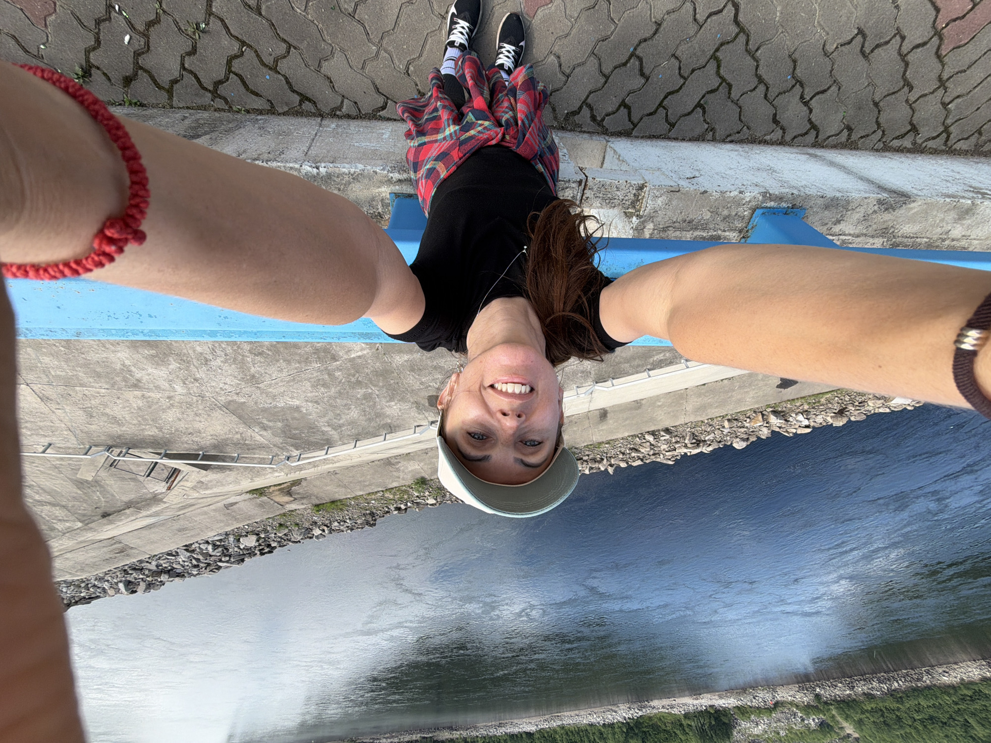 A woman stands at the edge of a wide river, possibly the Lena River near Talakan, Russia. She is taking a selfie, holding the camera out towards herself. The woman is wearing a black t-shirt, a plaid shirt tied around her waist, black leggings, white socks, and black and white athletic shoes. She also has a light-colored baseball cap on her head. A dark bracelet is on her left wrist and a red woven bracelet is on her right wrist. The river is dark blue and appears to be calm. Rocky banks line the river, and a concrete walkway with railings is visible in the foreground. The sky is overcast, reflecting on the water's surface.