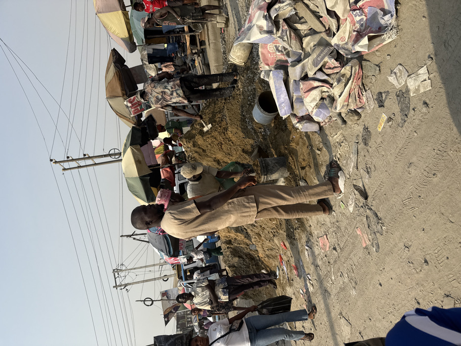 A bustling outdoor market scene with vendors under umbrellas lining the road. In the foreground, a man in a tan suit walks past a construction area where two workers mix cement. Bags of cement lie nearby. Overhead, power lines stretch across the sky. People stroll by, some carrying bags, adding to the busy atmosphere of the market.