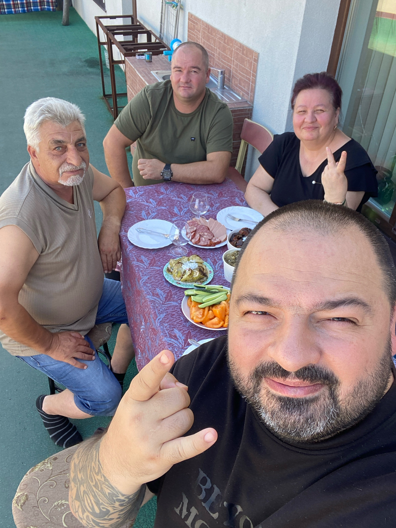The image depicts a group of four people, three men and one woman, gathered around a table outdoors. The setting appears to be a patio or backyard area, with a green-tiled floor and a white-tiled wall in the background. There's a sink and some metal shelving unit visible, suggesting an outdoor kitchen or preparation area. A table with a patterned tablecloth is set with plates, food (including what appears to be cold cuts, vegetables, and possibly some cooked dishes), and drinks. The people are casually dressed. One man in the foreground is taking a selfie, with his face close to the camera and his hand making a gesture. Another man is seated to his left, looking towards the camera. A third man is seated behind the table, also looking towards the camera. The woman is seated opposite him, making a peace sign with her fingers and smiling. The overall atmosphere suggests a casual gathering or meal. The specific location in Chișinău, Moldova, cannot be determined from the visual information provided.