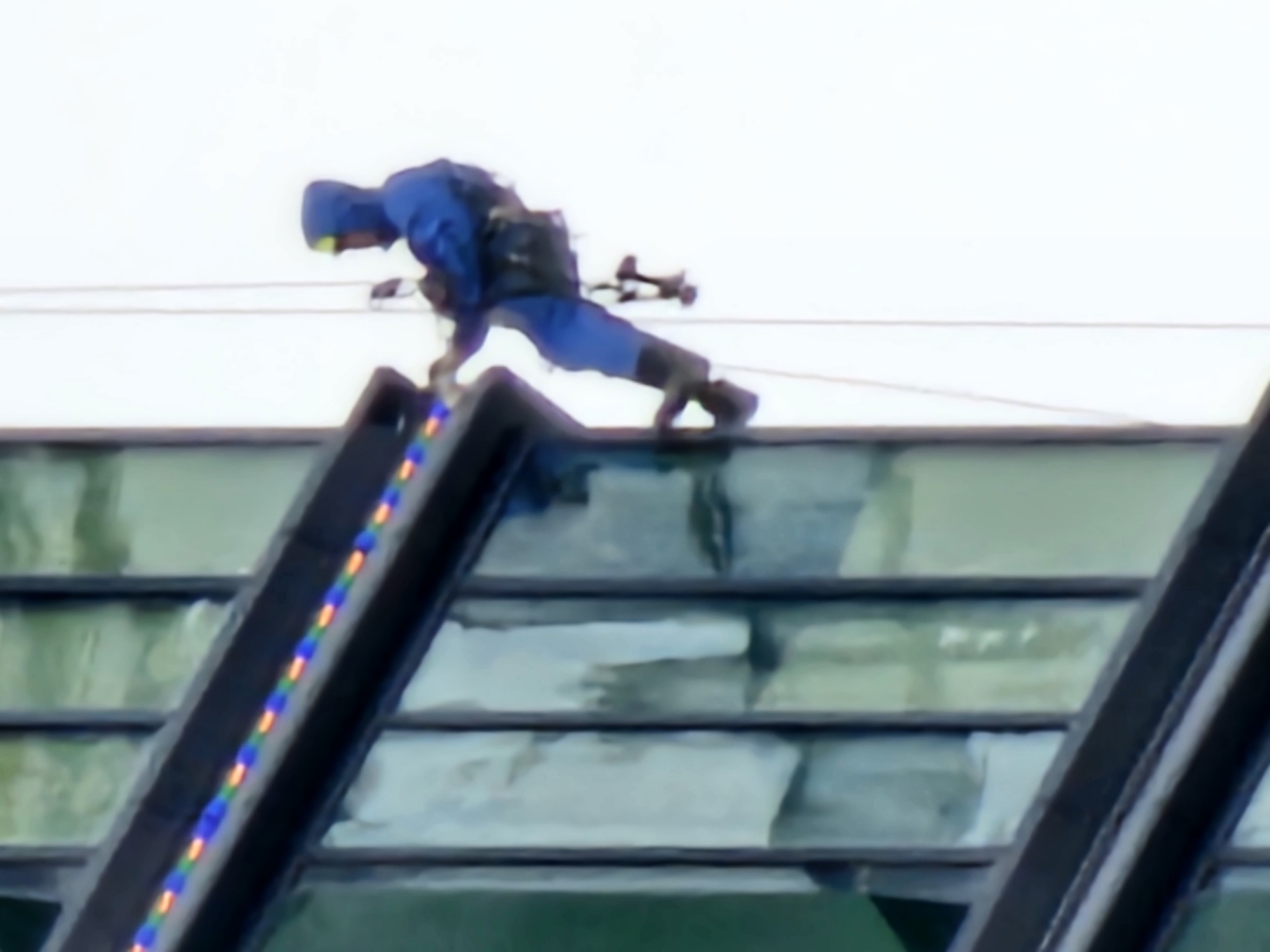 A person in a blue hooded outfit is rappelling down the side of a glass-paneled building. They are attached to a rope and harness, and appear to be working on the building's exterior. The building has dark framing around the glass panes, and a string of colorful lights is attached to the upper edge of the facade. The background is a bright, overcast sky. The image appears to be taken from a low angle, looking up at the person. It is not possible to determine the specific location within Moscow, Russia from the image alone.