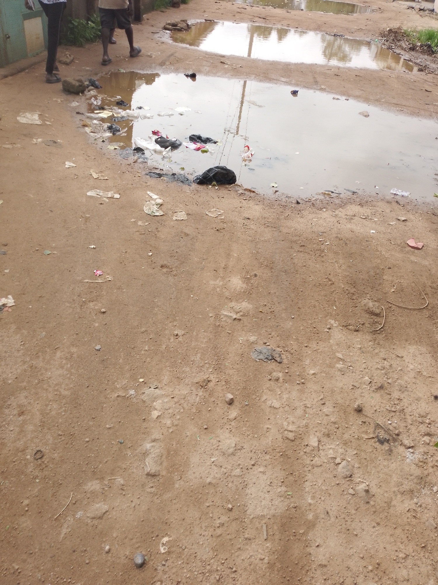 A high-angle medium shot shows a dirty, muddy ground with a large puddle of murky water in the upper portion of the frame. The ground is a light brown and appears to be dirt or sand. Scattered on the ground and in the puddle are various pieces of trash, including plastic bags, wrappers, and what appear to be discarded food items. In the upper left of the image, two individuals are partially visible, standing on the muddy ground. One person on the left is wearing dark pants and a white and black shirt, and the other person on the right is wearing dark shorts and sandals. The background shows more of the muddy ground and a very faint reflection in the puddle, possibly of trees or buildings, but it is unclear. The scene appears to be an outdoor area in a city, likely a street or alley, with a lack of proper waste disposal.