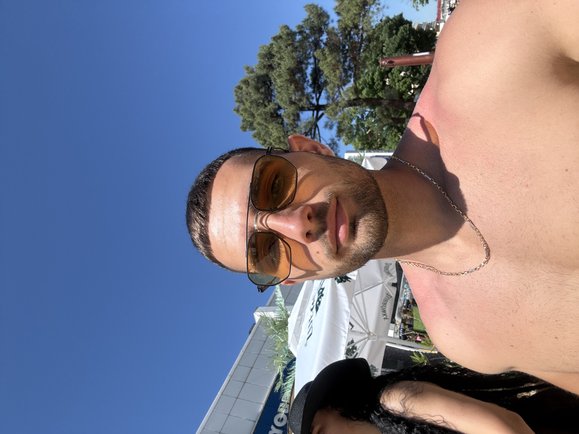 A close-up, selfie-style photograph captures a man with short, dark hair and a beard, wearing aviator-style sunglasses with orange lenses. He is shirtless, revealing tanned skin with a noticeable red sunburn on his right shoulder and upper back. A thin gold chain is visible around his neck. To his left, a person wearing a black wide-brimmed hat and a black top is partially visible. In the background, there is a white tent with visible text, a palm tree, and other foliage, suggesting an outdoor setting. The sky is clear and blue. The building in the far background suggests an urban or resort environment. Given the context of the question, this scene is likely in the city of Nesebar, Bulgaria, possibly near the coast or a beachside establishment.