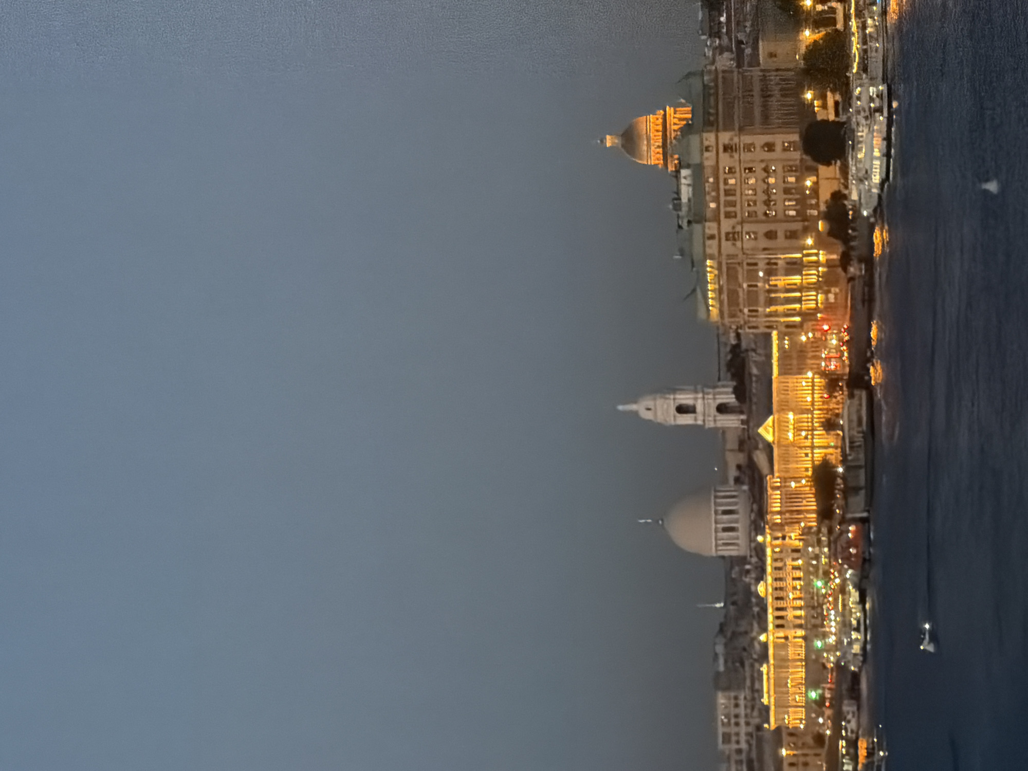 The image depicts a city skyline at dusk, likely in Saint Petersburg, Russia, given the architectural style. In the foreground, a body of water, possibly a river or canal, is visible with several boats of varying sizes illuminated by the city lights. The middle ground showcases a row of buildings lining the waterfront. These buildings are multi-story, with many windows lit up, creating a warm glow against the darkening sky. Distinct architectural features include domes and towers on some of the structures. One prominent building in the center has a large dome, and to its right, a tall, slender tower stands out. Further to the right, another large building features a prominent golden dome, illuminated by artificial lights, suggesting a significant landmark like a church or government building. The background consists of the sky, which is a deep blue-gray, indicating twilight or early evening. The overall atmosphere is serene and picturesque, highlighting the illuminated architecture of the city against the fading light.