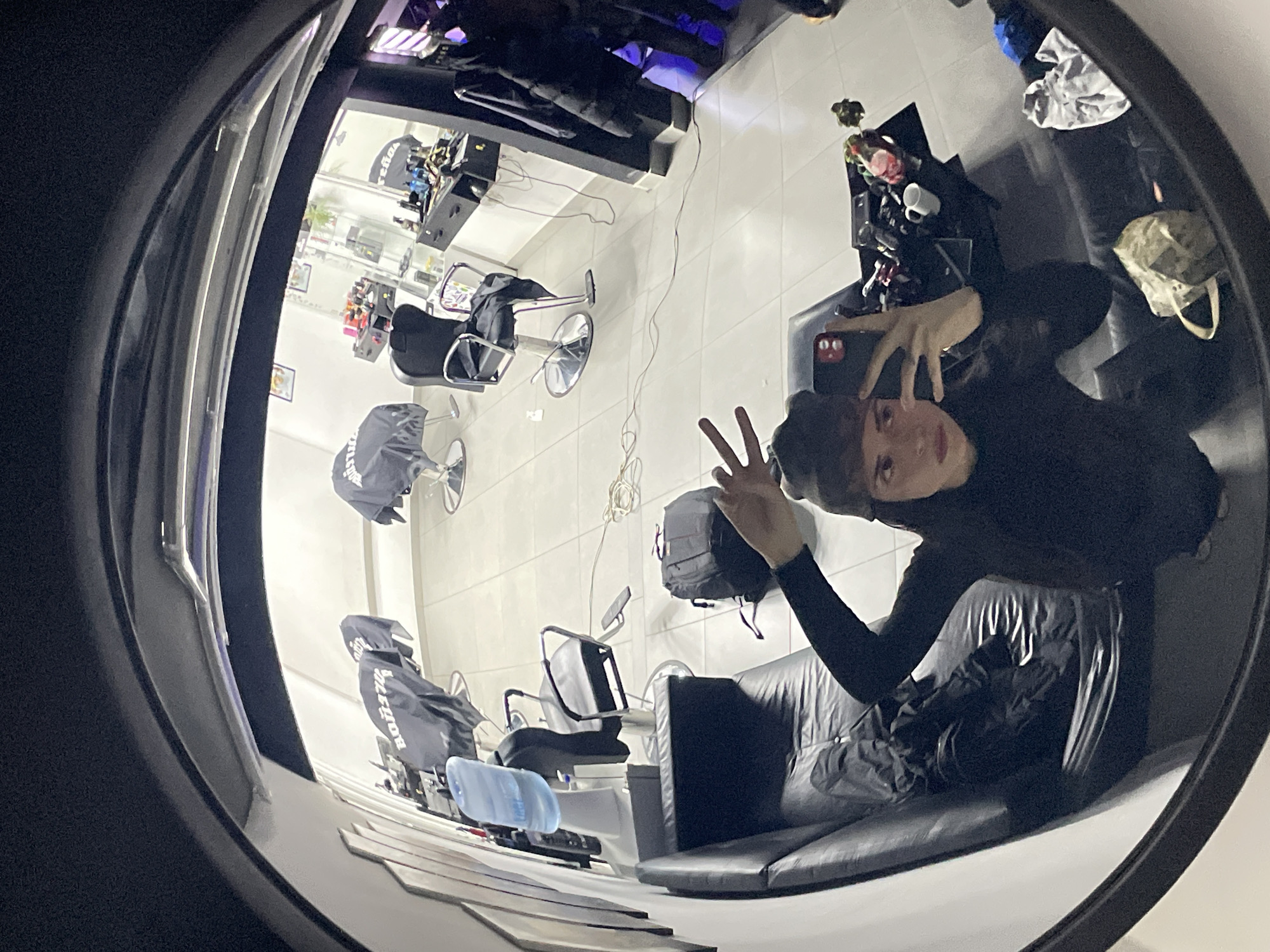 The image, taken with a fisheye lens, shows a man sitting on a black leather sofa in what appears to be a barbershop in Ushuaia, Argentina. The man is wearing a black shirt and has his hand raised, displaying a peace sign. The barbershop features multiple barber chairs with protective covers, a workstation with various tools and products, and a tiled floor. The reflection in the mirror suggests the shop has a modern interior with good lighting. A backpack is near the sofa. A water bottle is near the barber chairs. Outside the window, the store appears to be situated in a commercial area.