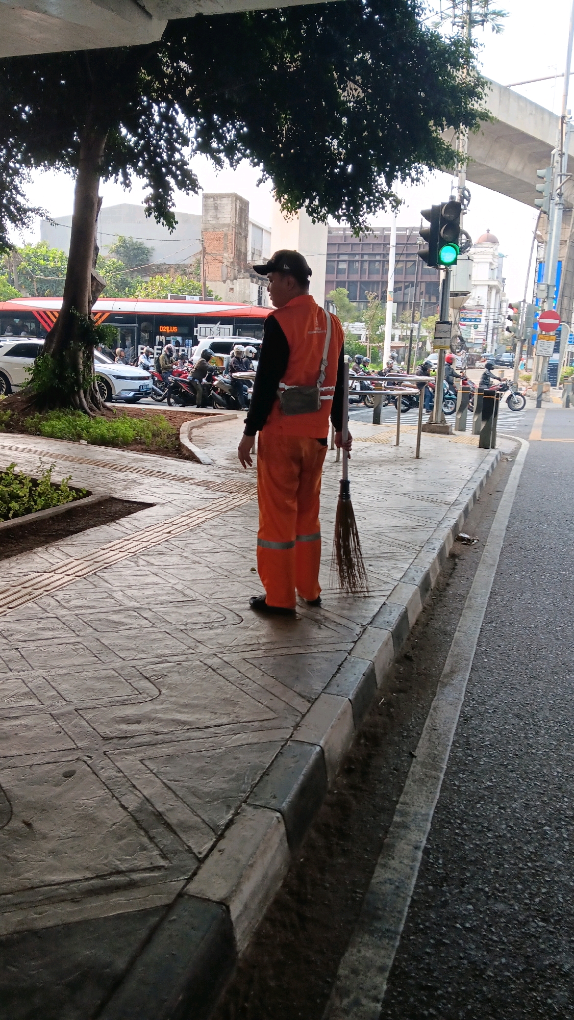 A street cleaner stands on a sidewalk in South Jakarta, Indonesia, holding a broom. The street cleaner is wearing an orange uniform with reflective stripes and a black cap. In the background, there is a street with traffic, including cars and motorcycles. There is also a bus and a traffic light that is green. The sidewalk is made of patterned concrete and has a curb. There is a tree with dark green leaves to the left of the image, and an elevated road structure in the background.