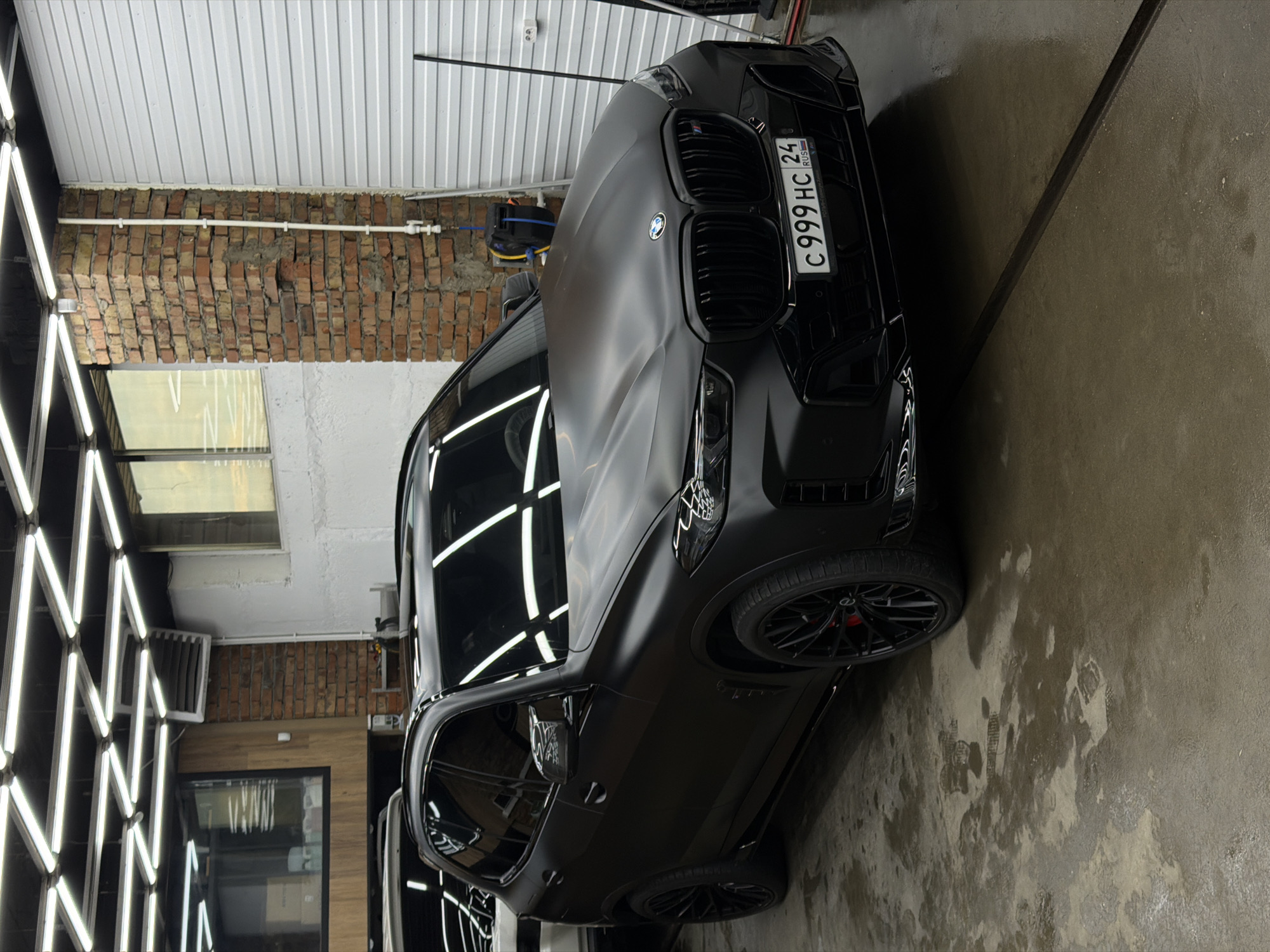 The image depicts a matte black BMW X6 M Competition in a car detailing bay. The vehicle has a Russian license plate, "C 999 HC 24 RUS," indicating it is registered in the Krasnoyarsk region. The bay has a brick wall and a corrugated metal ceiling. Artificial lights provide illumination. The floor is wet, showing shoe prints. The car is parked close to the side wall, with the front end closer to the camera.