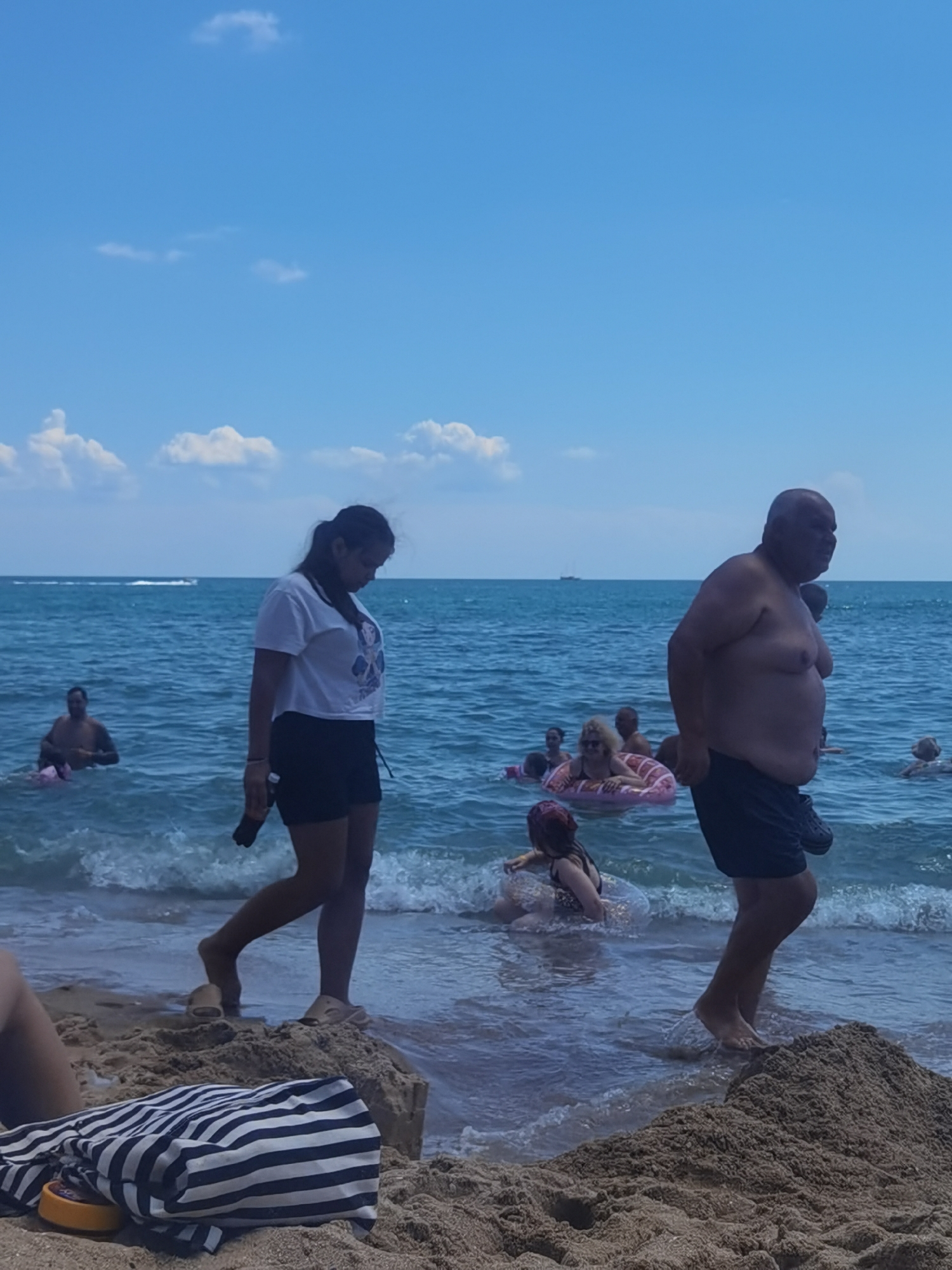 The image depicts a sunny day at a beach, likely in Varna, Bulgaria, with people enjoying the sea. In the foreground, on the sandy shore, lies a black and white striped towel with an orange object beside it. A person's legs are visible on the sand near the water's edge. In the shallow water, a woman in a white t-shirt and black shorts walks towards the viewer, holding a dark object in her hand. Further out in the sea, several individuals are swimming and relaxing. One man is partially submerged in the water. In the middle ground, a group of people are floating on inflatable rings, with one woman appearing to be adjusting her ring. In the background, the blue sea stretches out under a clear sky with scattered white clouds. A distant boat is visible on the horizon. The overall scene is one of leisure and recreation at the seaside.