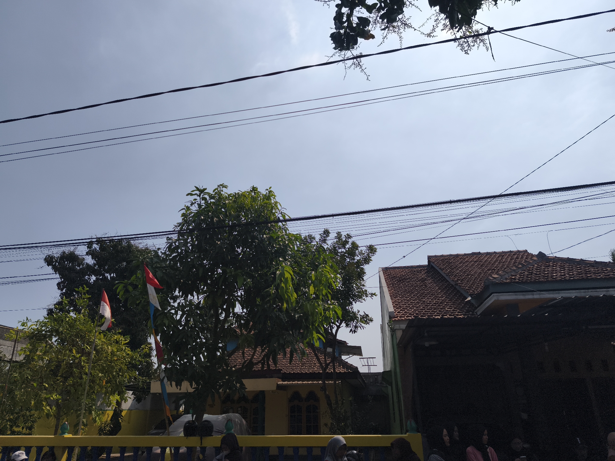 The image captures a street scene in Kutoarjo, Indonesia, with several houses visible. In the foreground, a yellow fence separates the viewer from a group of people, some wearing headscarves, standing or sitting. Behind the fence, trees with lush green foliage are prominent, with two Indonesian flags flying from poles amidst the leaves. The flags, red and white, are partially unfurled.
Behind the trees, a single-story yellow house with a tiled brown roof is visible. To the right, another house with a similar tiled brown roof is partially obscured by a dark, shaded area that appears to be an overhang or a covered structure. The sky above is clear and pale blue, with numerous electrical wires crisscrossing the frame, indicating an urban or semi-urban setting. The overall impression is a typical residential area on a bright day.